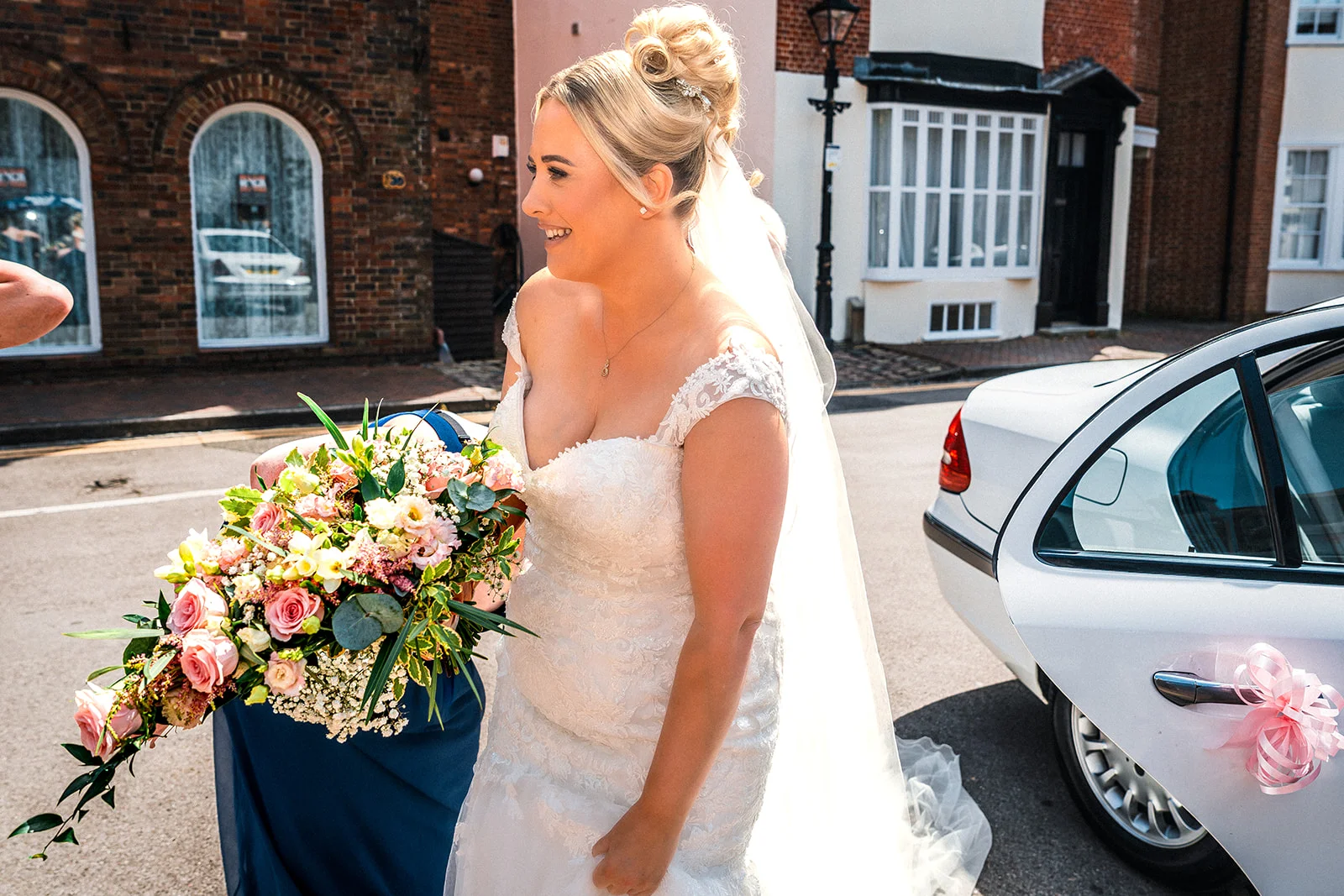 A smiling bride holding a cascading floral bouquet steps out onto a sunny street near a classic white wedding car.