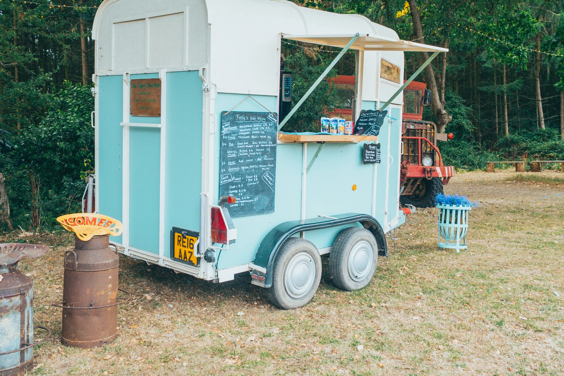 A converted light blue horsebox trailer serves as an outdoor mobile bar parked on a grassy woodland clearing during a festival-style wedding.