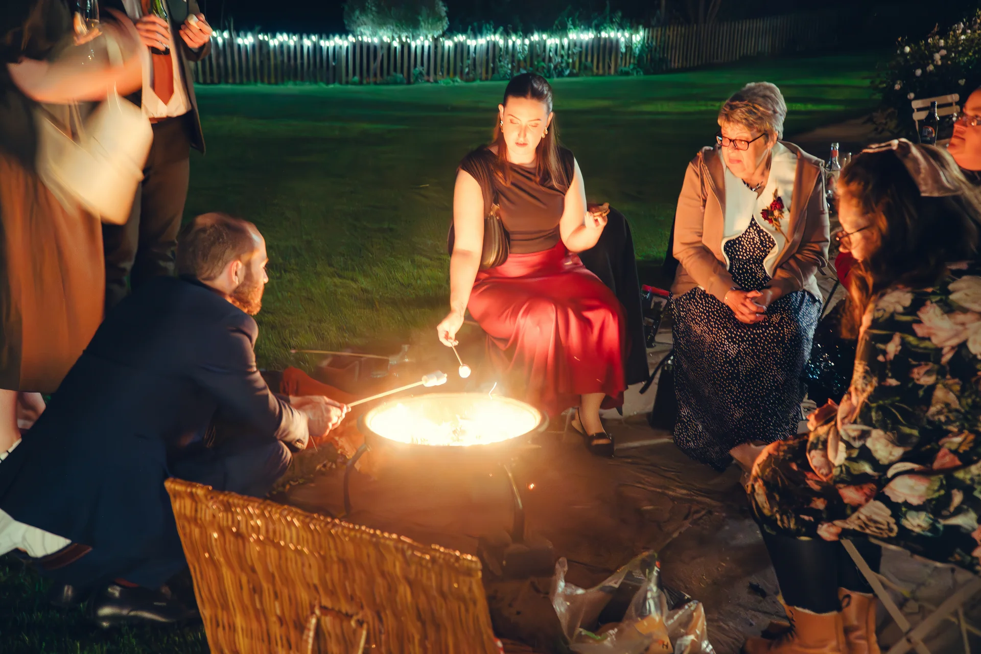 Wedding guests gather around a glowing firepit outdoors at night, toasting marshmallows and chatting by the lawn.