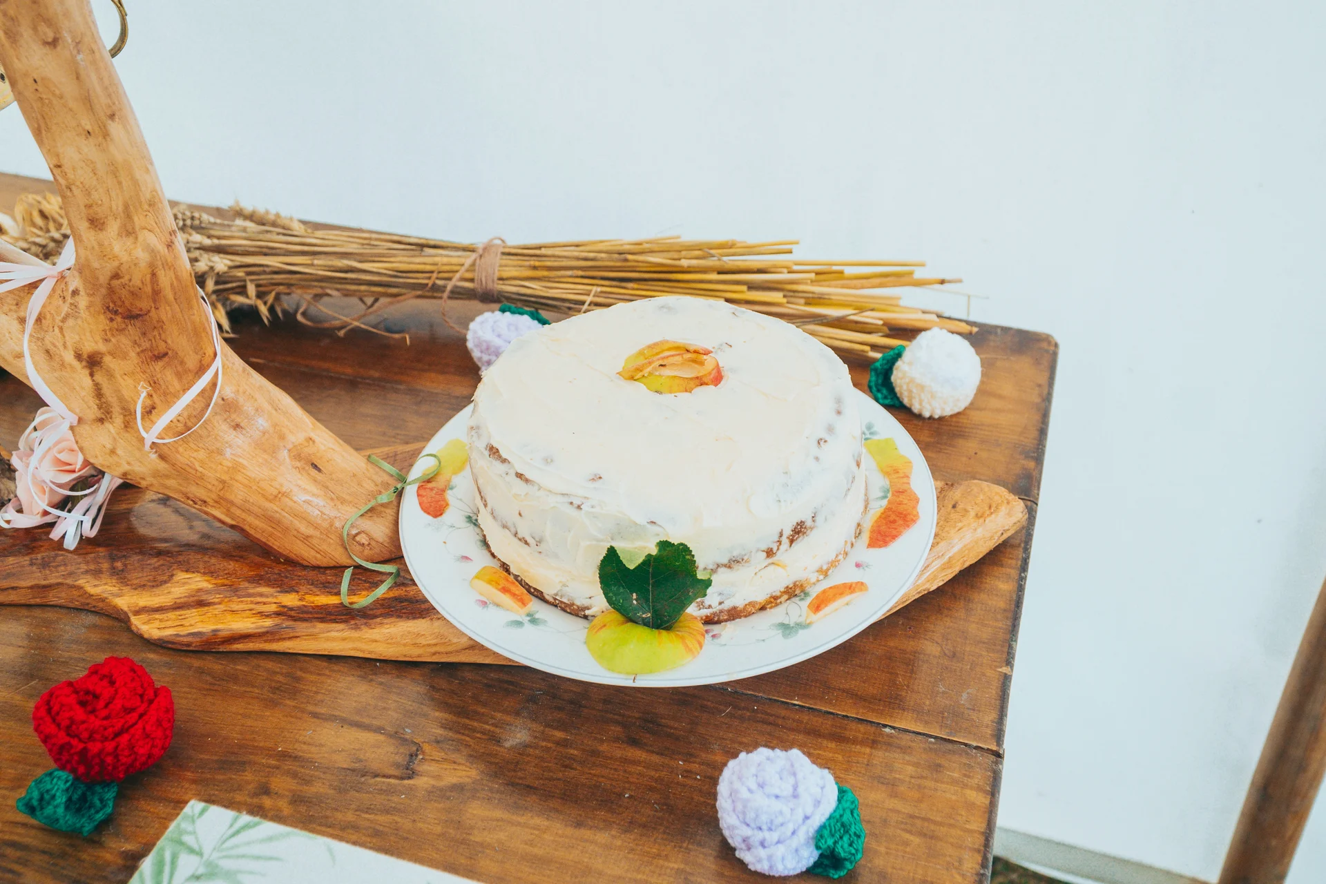 A rustic semi-naked single-tier wedding cake decorated with sliced fruit and greenery sits on a wooden table alongside dried wheat and crocheted floral details.