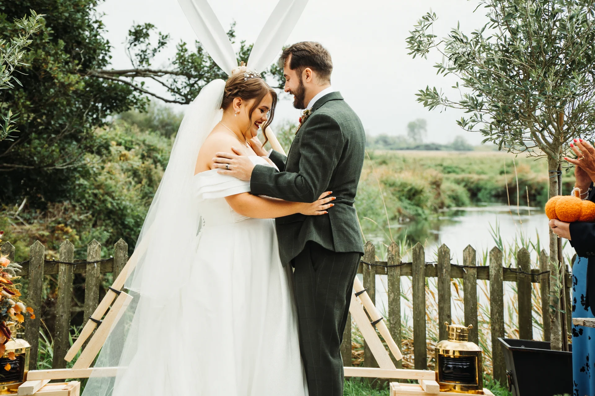 A smiling bride and groom embrace affectionately during an outdoor wedding ceremony set against a picturesque riverside landscape.