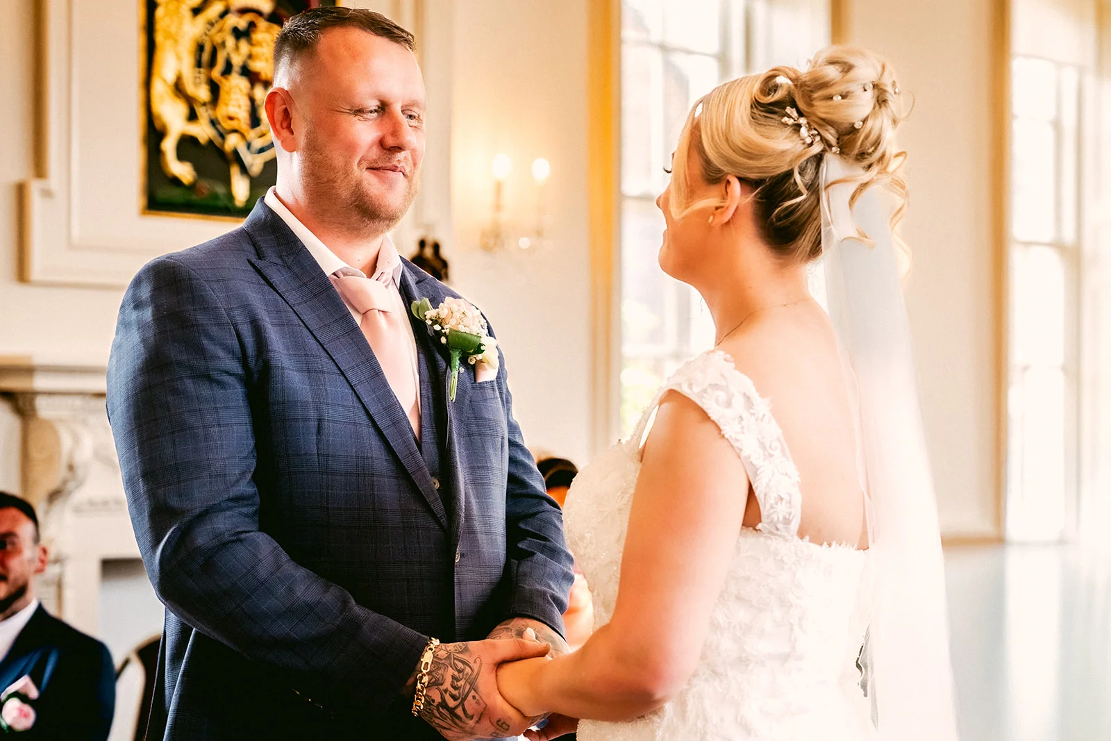 A smiling groom in a blue checked suit holds hands and exchanges loving glances with his bride during their indoor wedding ceremony.