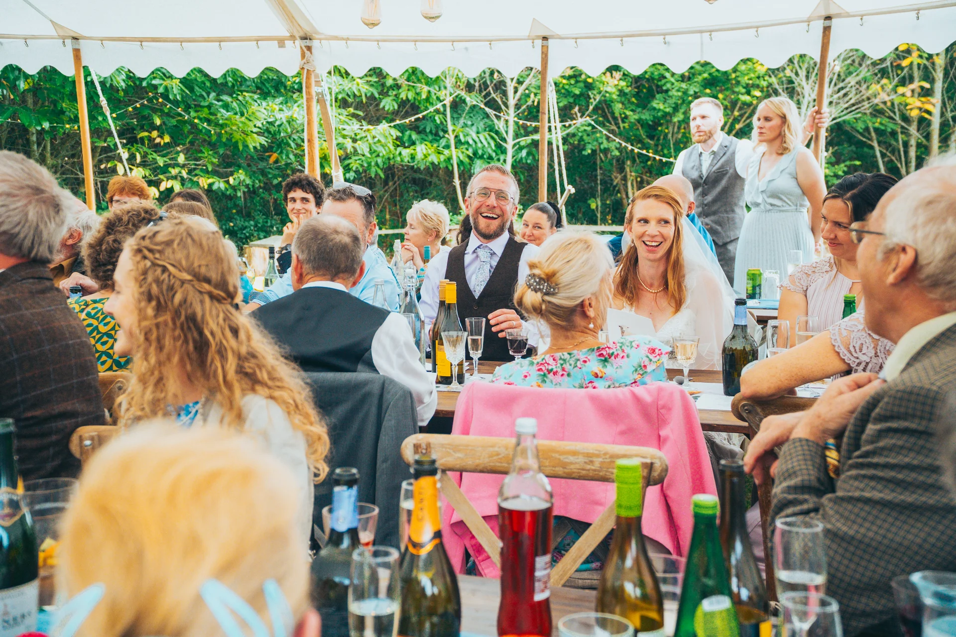 Happy wedding guests seated at long banquet tables inside a marquee sharing a joyful moment during speeches.