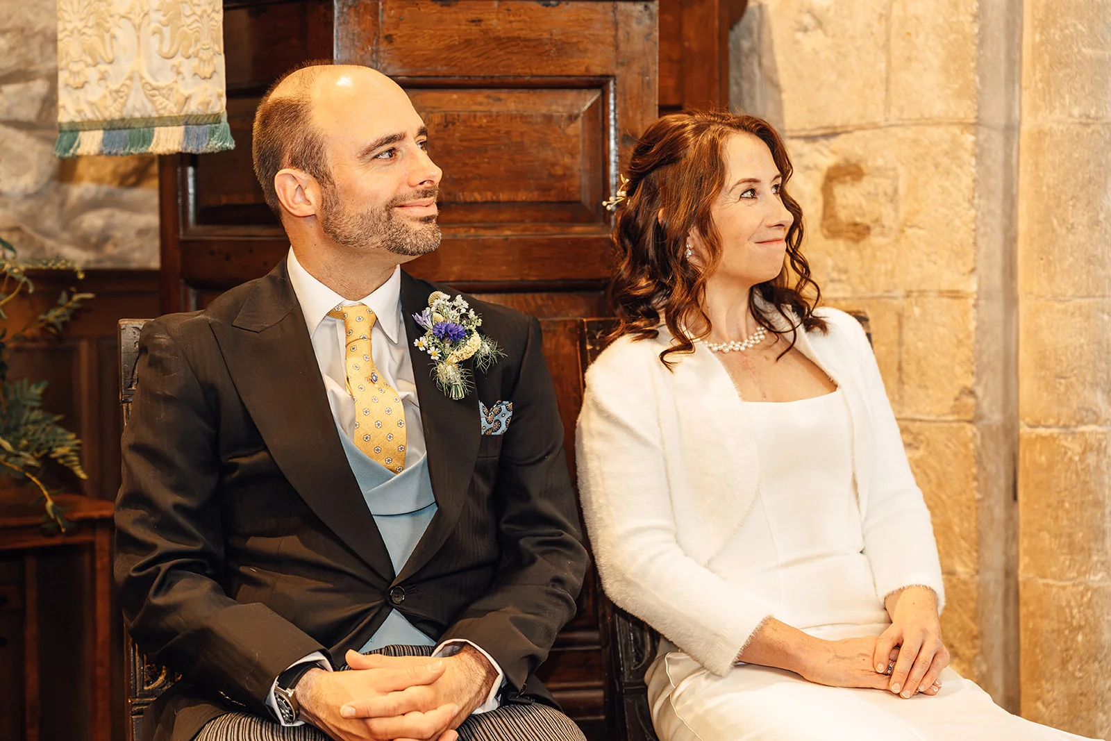 A mature bride and groom sit side by side during their church wedding ceremony, both looking thoughtfully towards the side.