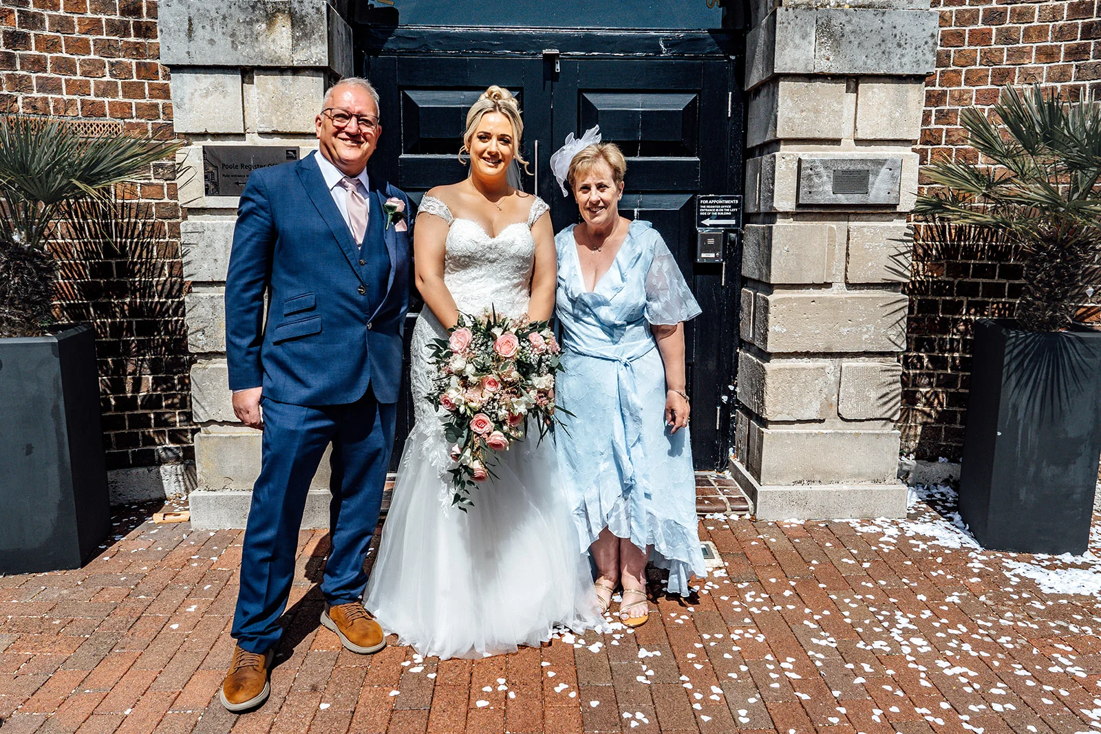 A smiling bride stands outdoors in her wedding dress holding a cascading bouquet, flanked by her parents outside a registry office entrance.