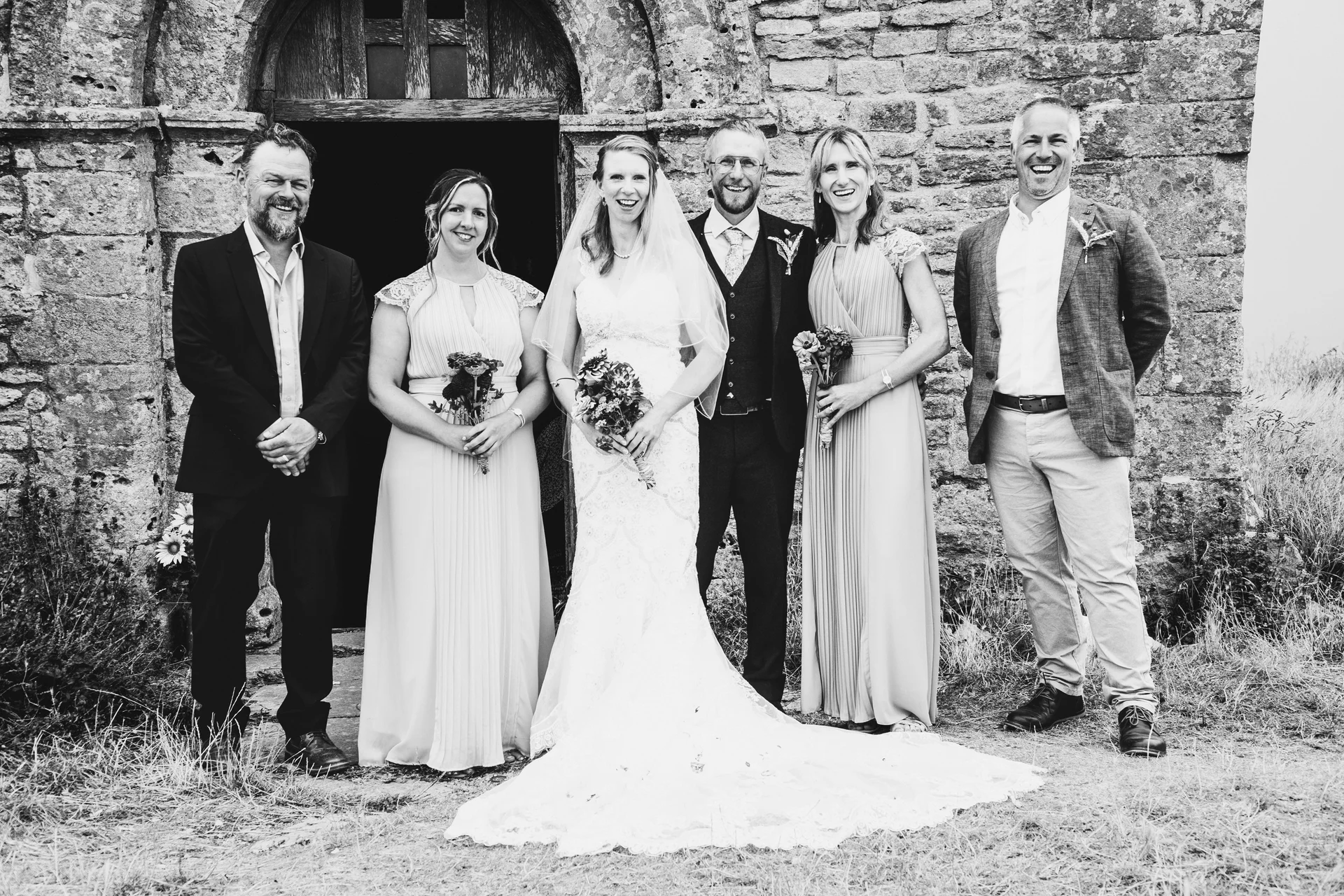 A newlywed couple and their wedding party smile outside the arched wooden doorway of a rustic stone church in this black and white group portrait.