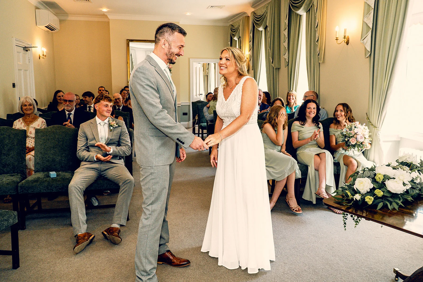 A newlywed couple holds hands and smiles joyfully during their indoor civil ceremony while seated guests clap and celebrate.