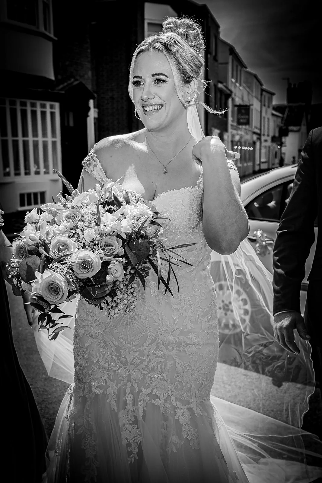 A joyful bride in a lace off-the-shoulder gown laughs outdoors on a street, holding a large floral bouquet and wearing her hair in a high updo.