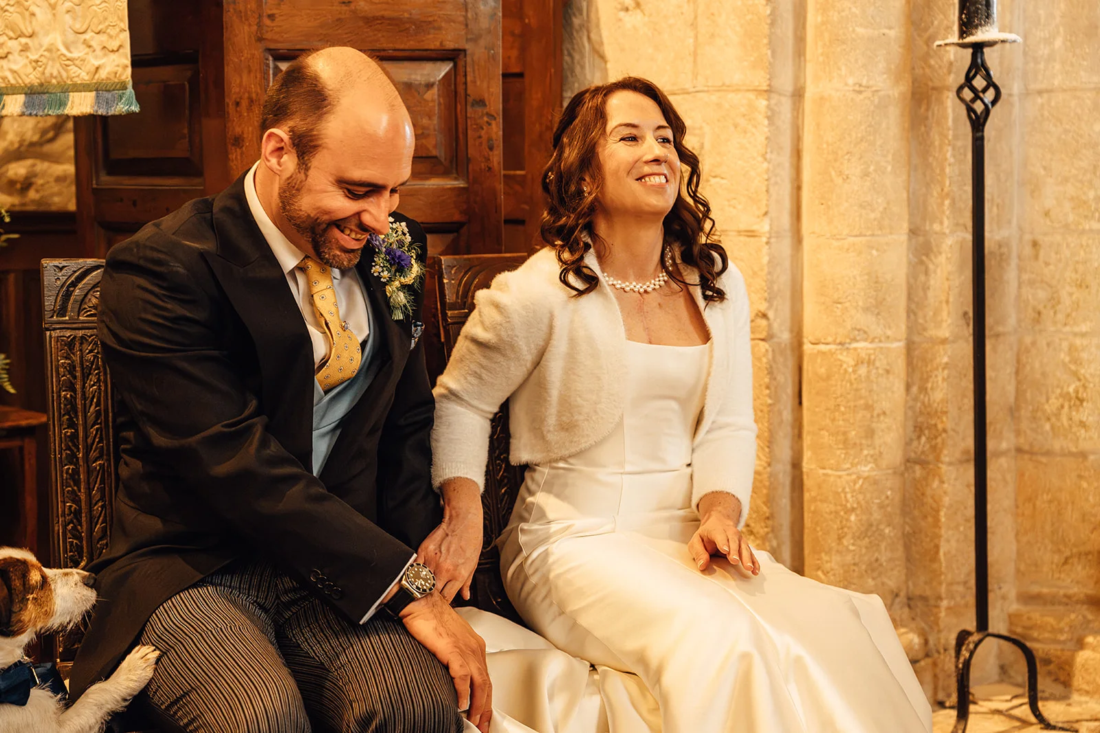 A mature bride and groom sit together inside a stone church, laughing joyfully as the groom looks down with a smile.