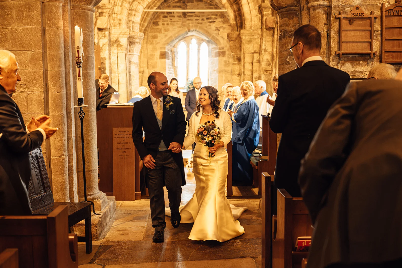 The newly married couple walk hand in hand down the stone aisle of a historic church while smiling guests applaud their exit.