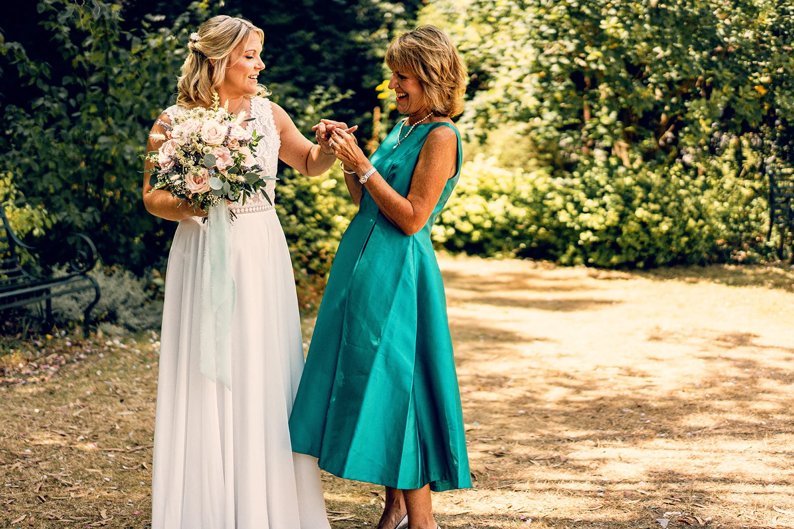A smiling bride holding a floral bouquet shares a tender, laughing moment in a sunny garden with her mother wearing an emerald green dress.
