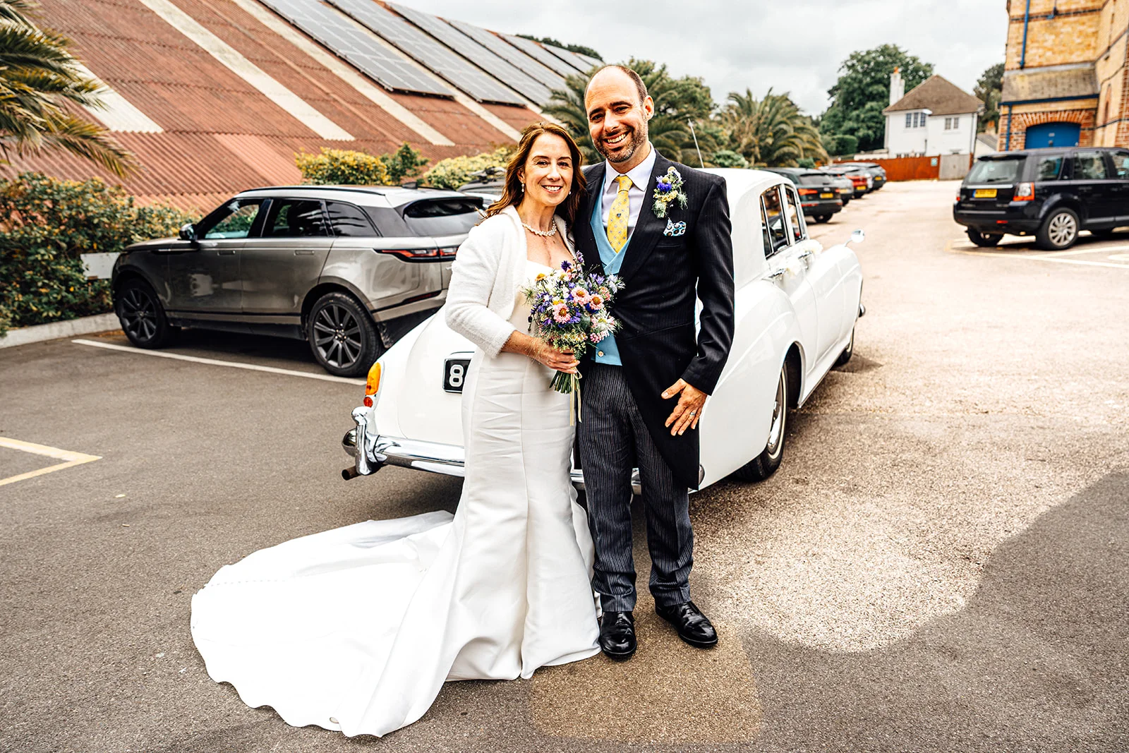 A smiling bride in a white satin trumpet gown with a chapel train and a groom in a morning suit pose outdoors next to a vintage white wedding car.