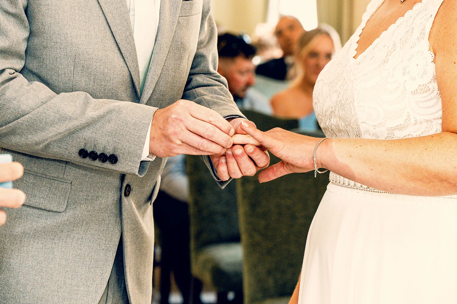 A groom in a grey suit gently places a wedding ring onto the finger of a bride wearing a lace dress during their ceremony.