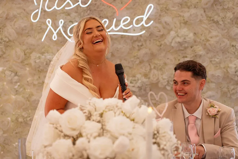 A joyful bride laughs heartily into a microphone while delivering her speech at the top table, with the groom smiling lovingly beside her in front of a white floral wall and a neon sign.