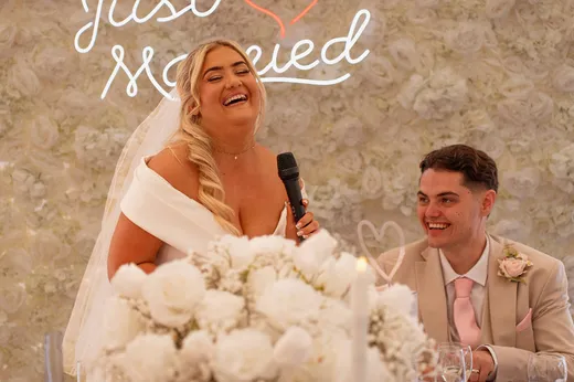 A joyful bride laughs heartily into a microphone while delivering her speech at the top table, with the groom smiling lovingly beside her in front of a white floral wall and a neon sign.
