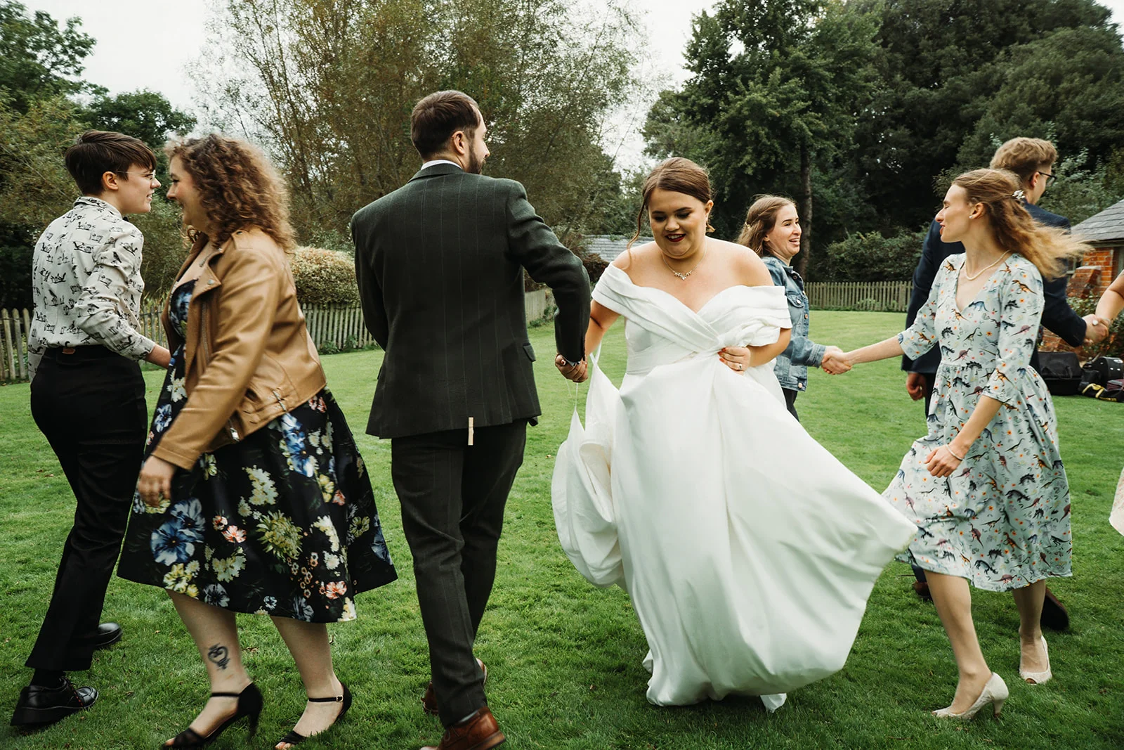 A newlywed couple and their wedding party joyfully dance and walk across a lush green lawn in a vibrant, candid outdoor setting.