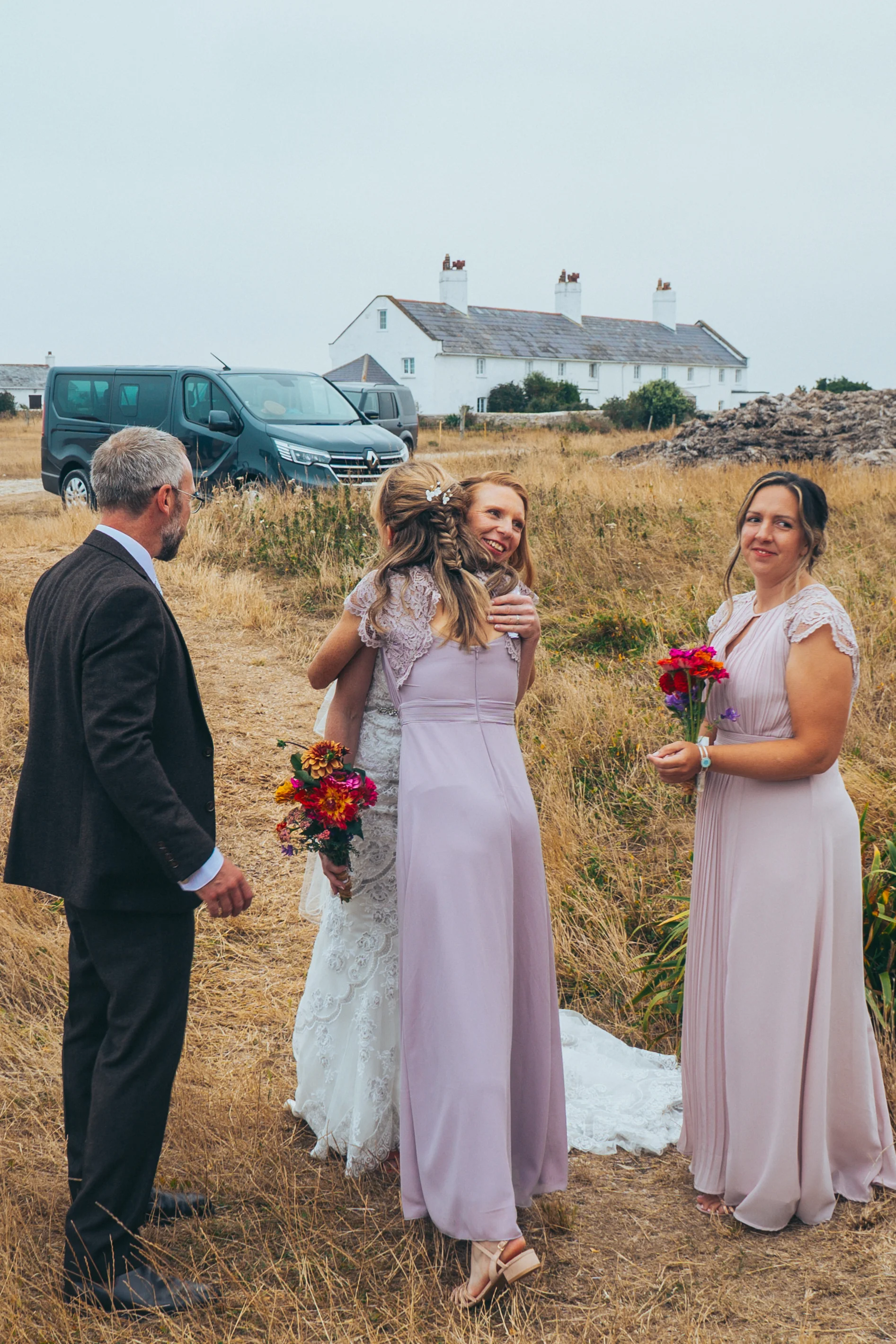 A bride in a lace wedding dress embraces her bridesmaids in pink dresses during an outdoor rural celebration under an overcast sky.