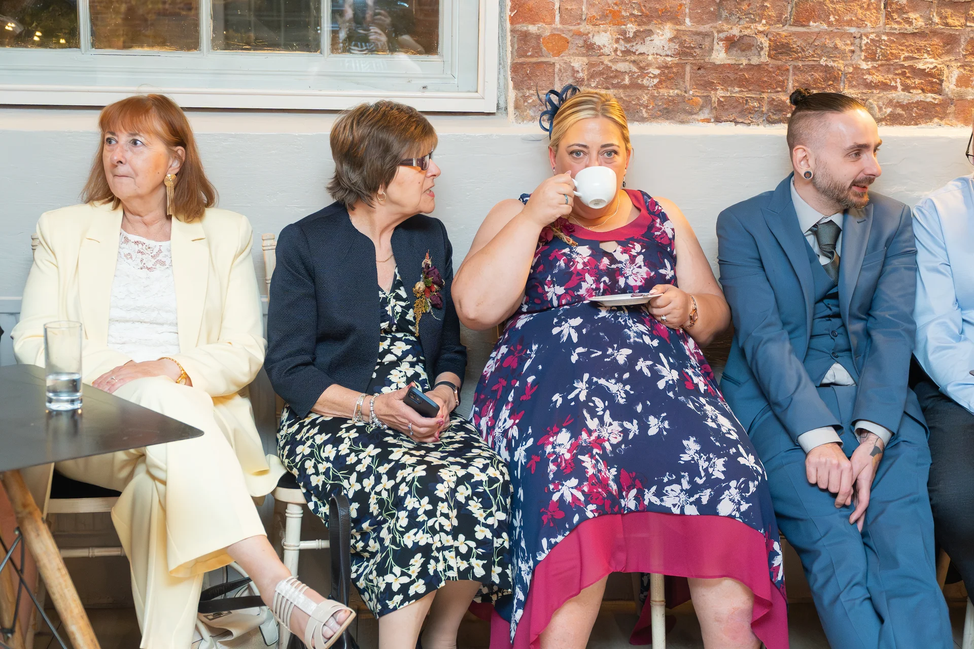 Wedding guests sit together indoors against a brick wall, with one woman drinking tea from a white cup and saucer.