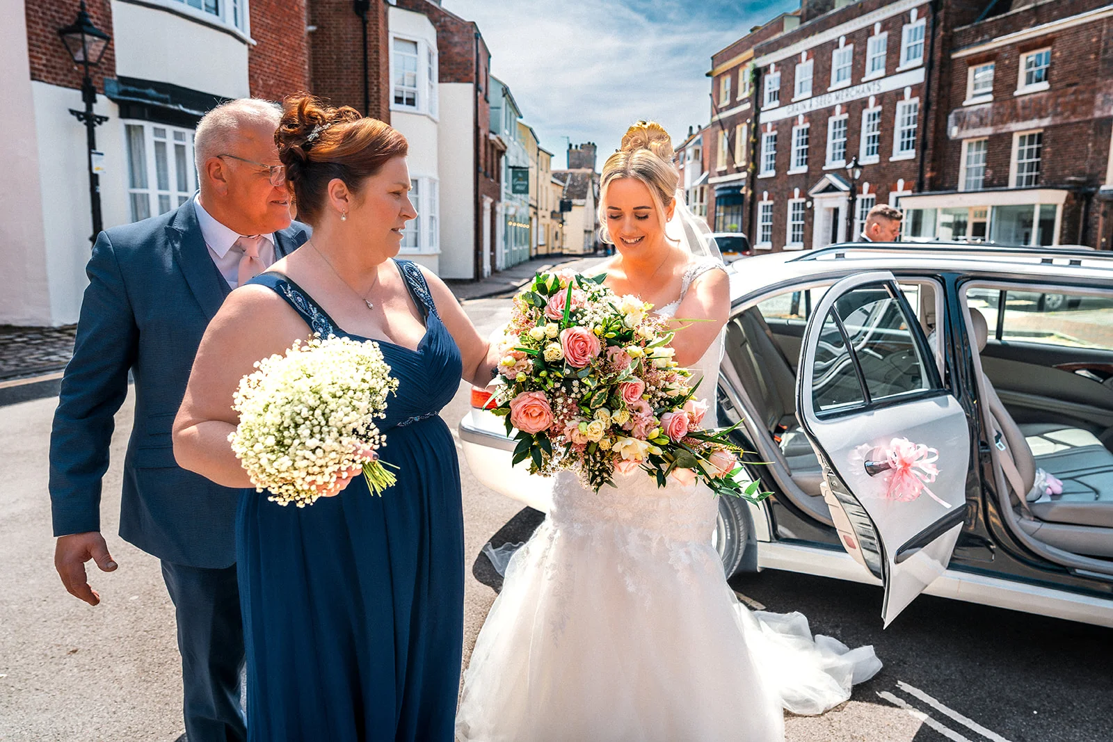 A radiant bride holding a large floral bouquet steps out of a wedding car on a sunny street, accompanied by her father and a bridesmaid.