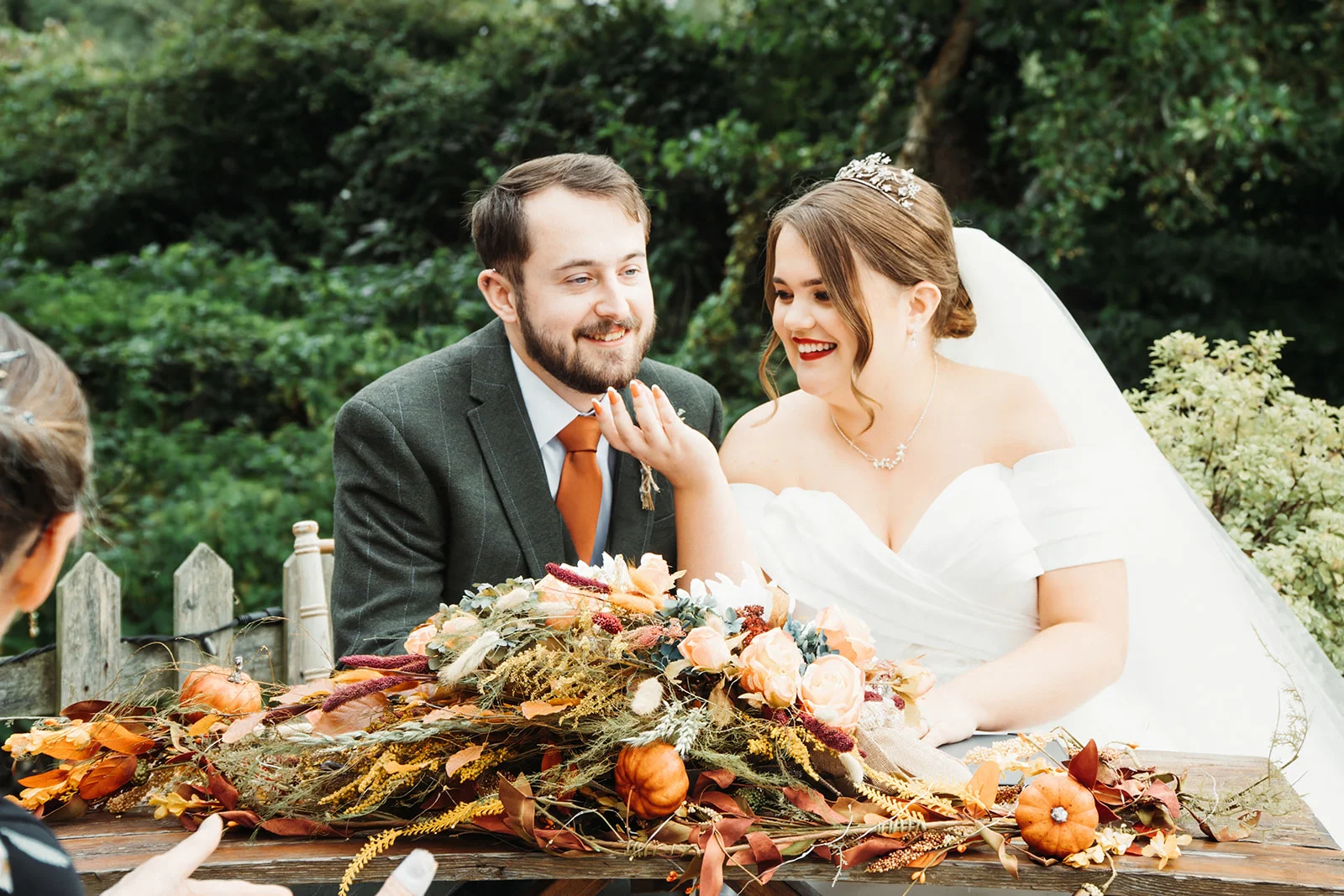 A joyful newlywed couple seated outdoors at a rustic wooden table decorated with autumn leaves and mini pumpkins sharing a lighthearted moment during their wedding reception.