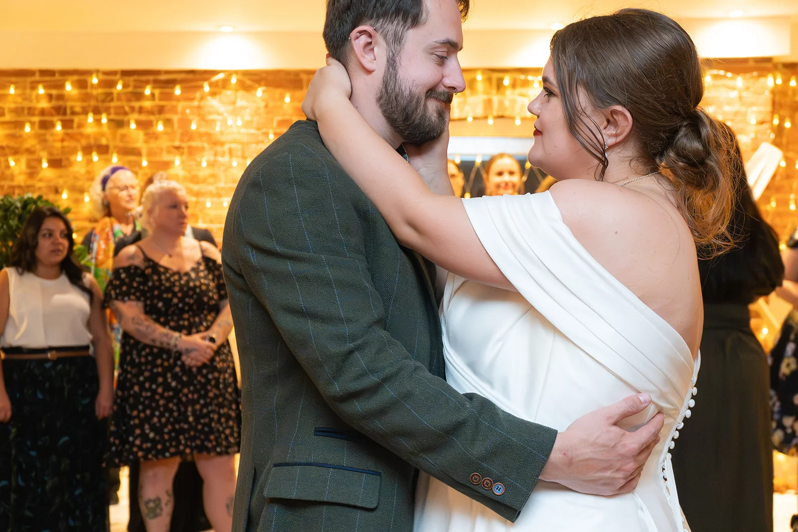 A bride and groom share a tender moment dancing together during their evening reception against a backdrop of exposed brick and glowing string lights.