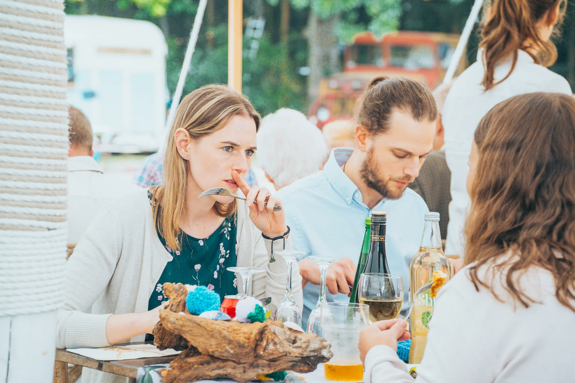Guests enjoy a relaxed outdoor wedding breakfast, seated together at a rustic wooden table with drinks and casual dining.