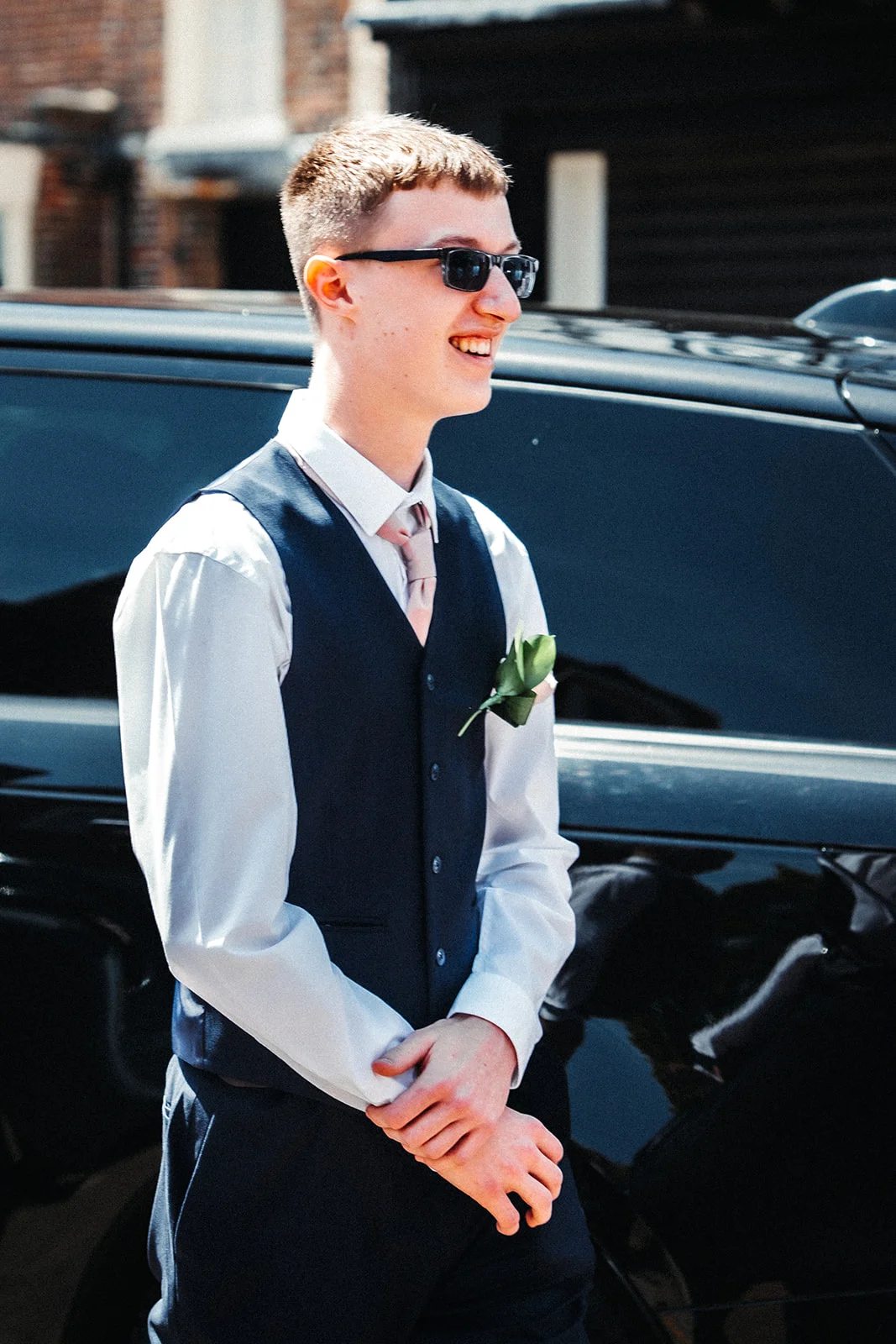 A smiling young groom wearing a dark waistcoat, white shirt, and sunglasses stands near a shiny black car on his wedding day.