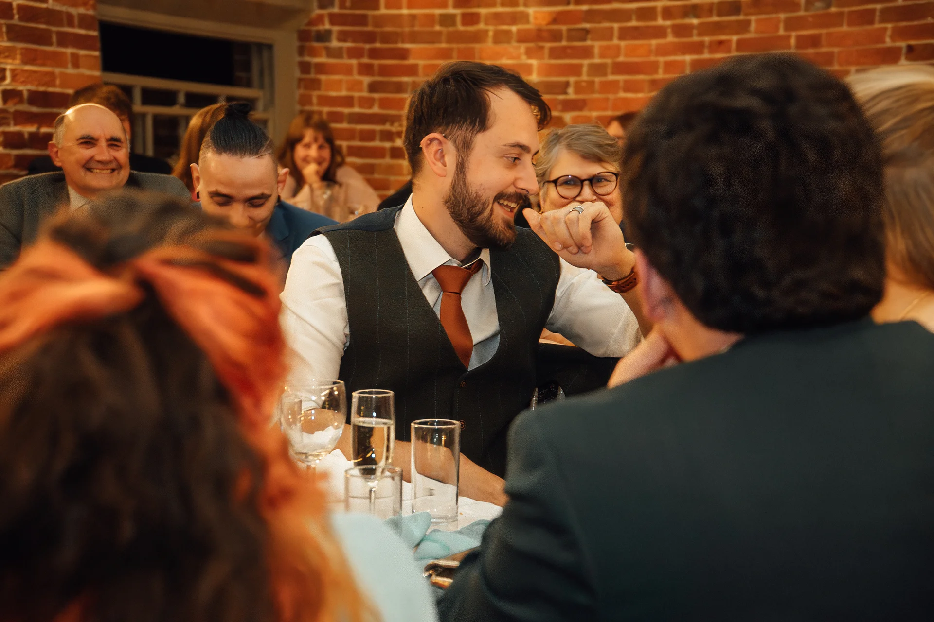 A smiling groom in a waistcoat speaks during the wedding breakfast while guests laugh and listen attentively in a room with exposed brick walls.