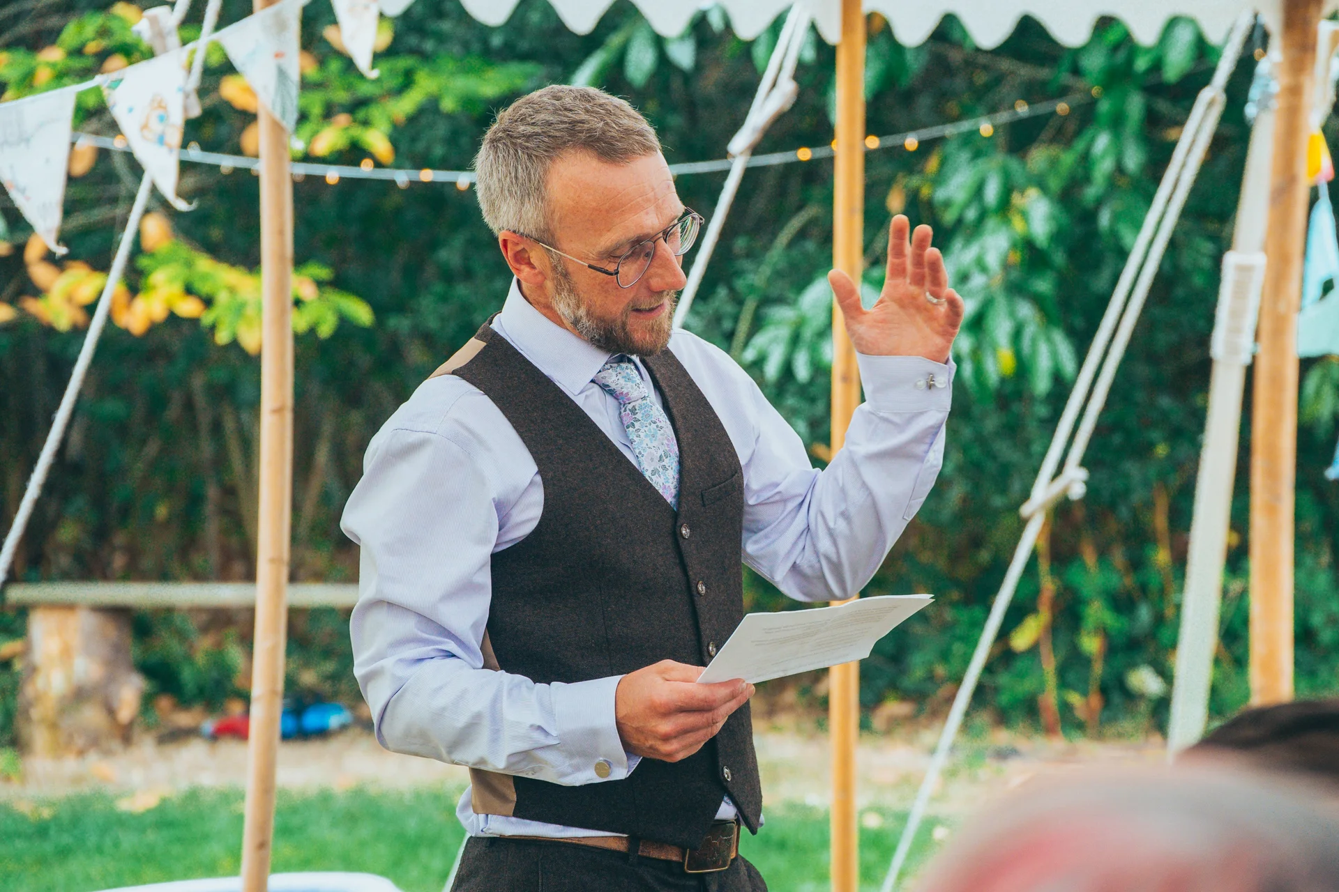 A groom wearing a brown tweed waistcoat and patterned tie delivers a speech inside a marquee, gesturing animatedly while holding notes.