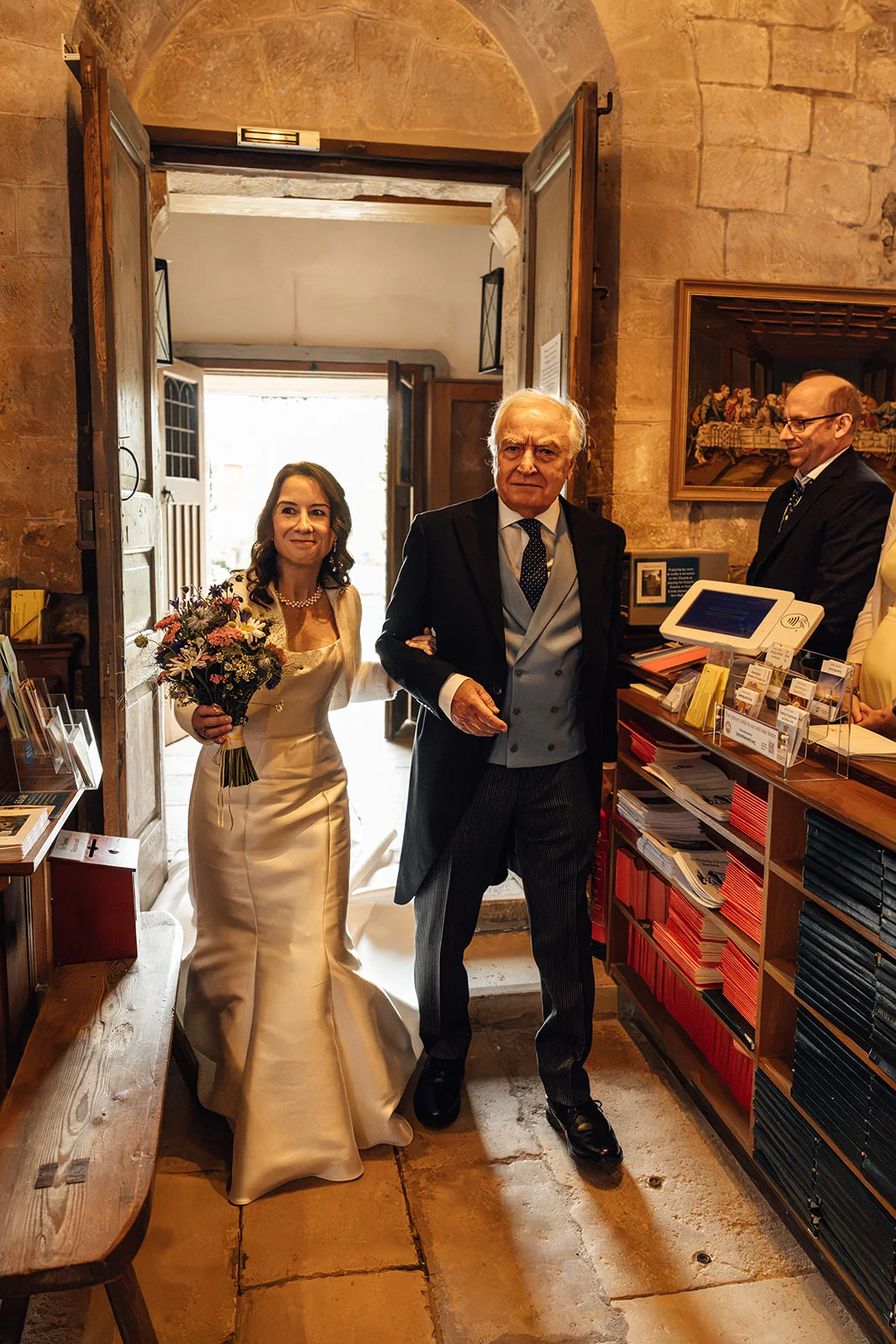 A smiling bride holding a wild-style bouquet walks arm in arm with her father through a stone church doorway.