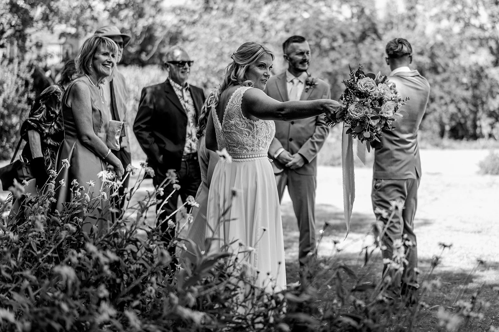 A smiling bride in a lace-detailed gown gestures playfully with her bouquet during an outdoor wedding ceremony while guests watch and laugh.