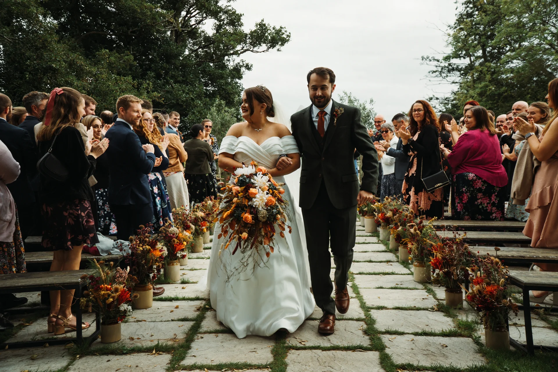 Newlywed bride and groom happily walk down the stone aisle together amidst applauding guests, holding a large autumnal bouquet filled with burnt orange and dried floral elements.