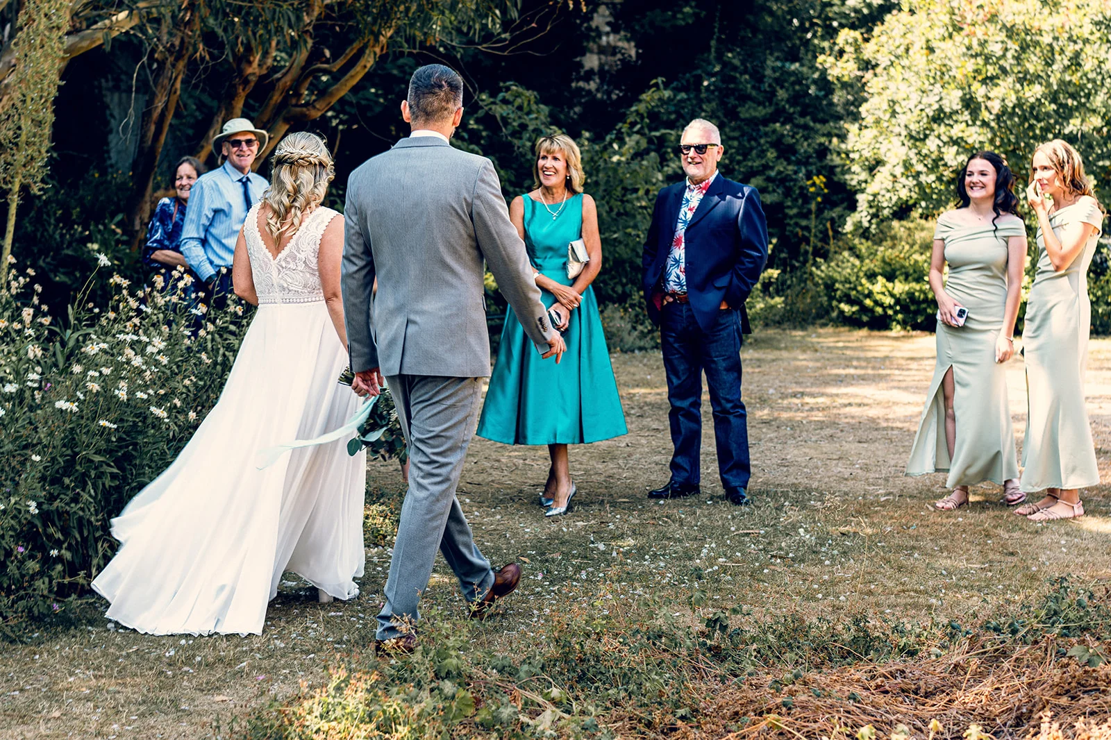 A bride in an a line gown and a groom in a grey suit walk through a sunlit garden past family members and guests.