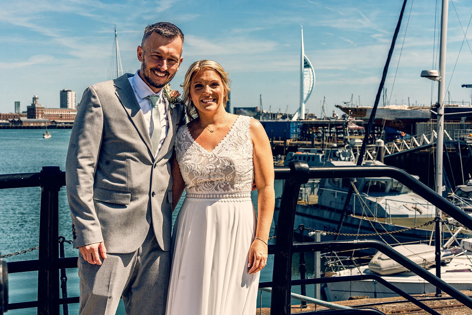 A smiling bride and groom pose together by a scenic harbour with boats and a tall white tower in the background on a sunny day.