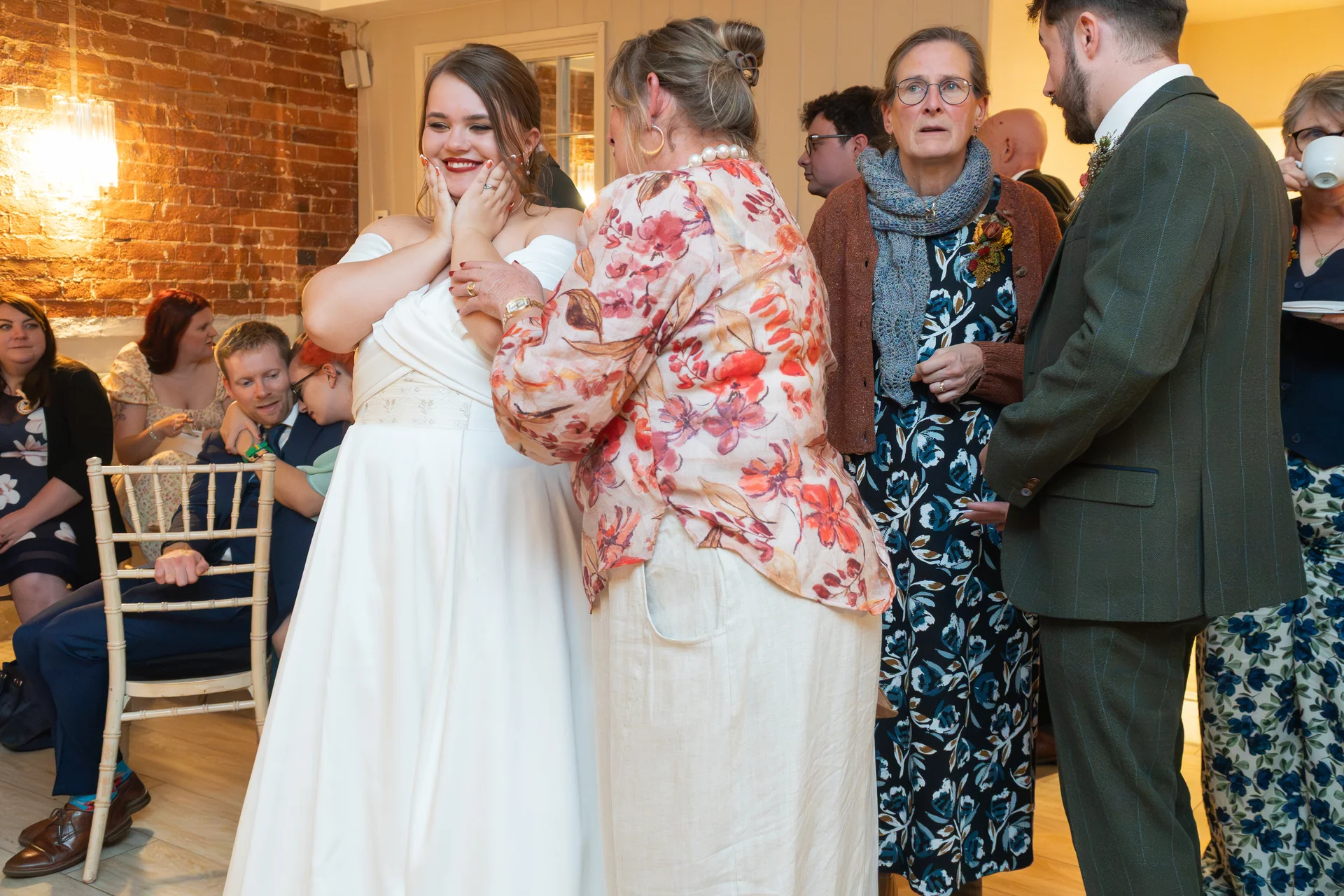 A joyful bride in a white off-the-shoulder gown laughs with her hands to her face while guests mingle around her indoors.