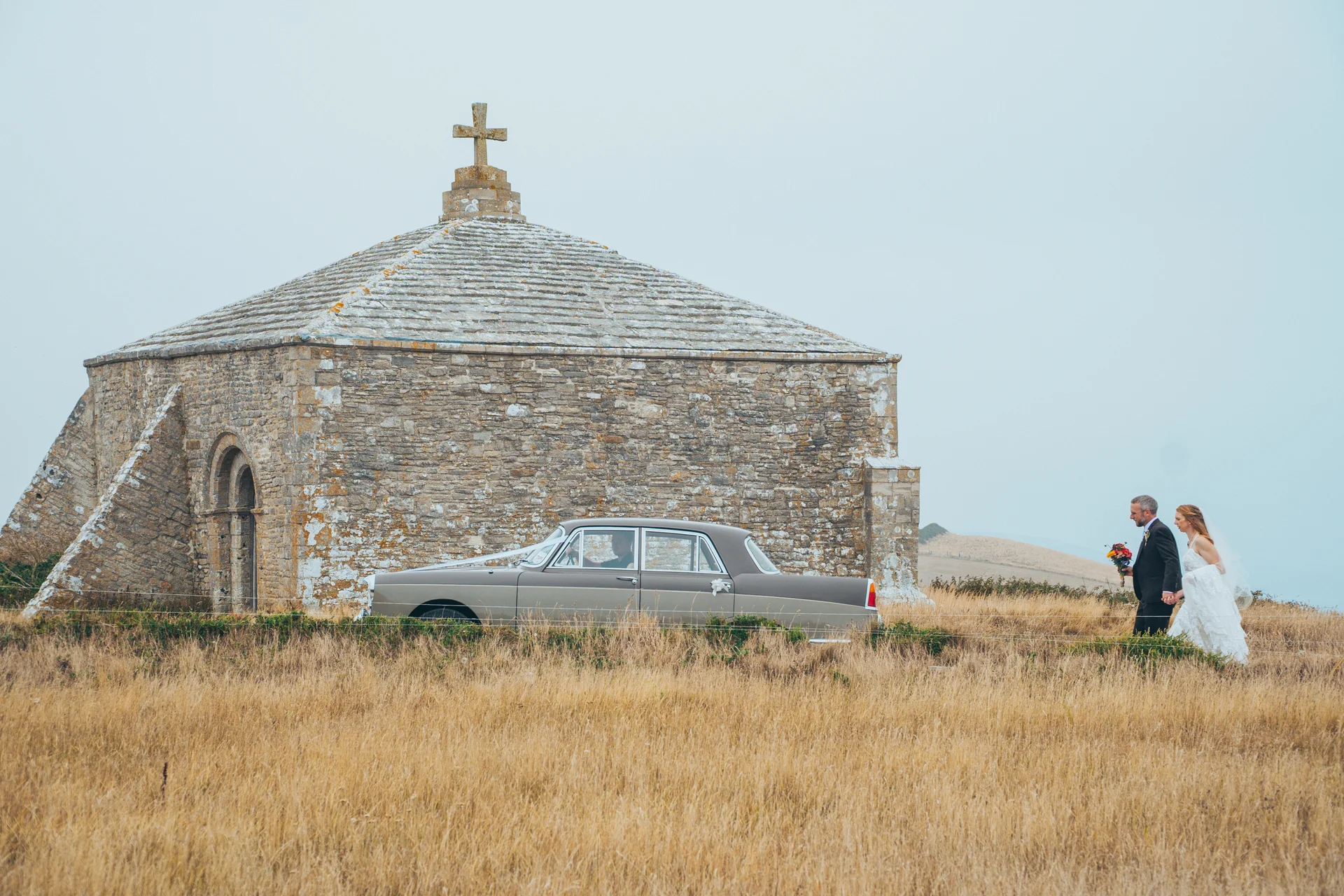 A newlywed couple walks hand in hand across a coastal meadow next to a classic car and a historic stone chapel under an overcast sky.