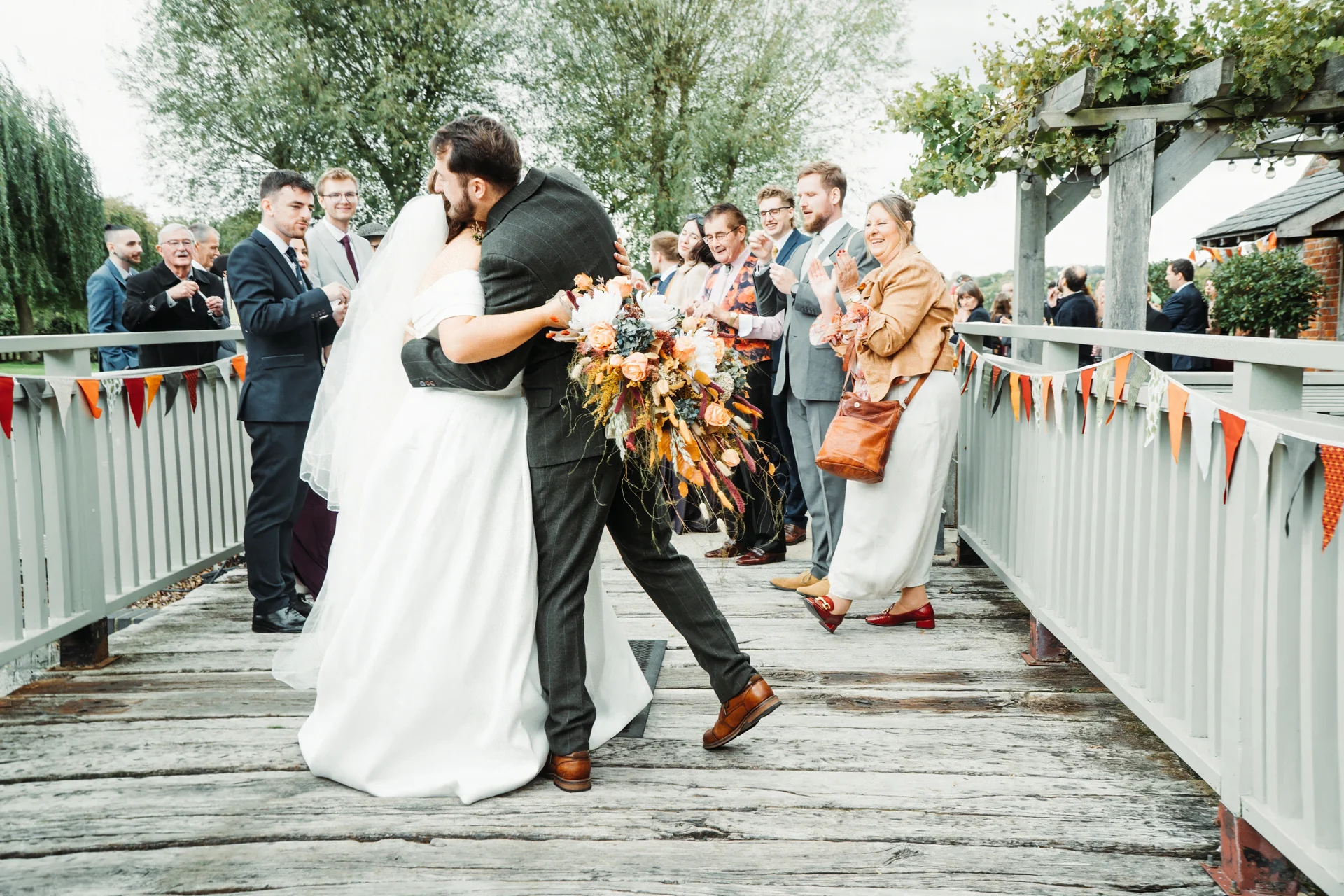 A newlywed couple embraces and shares a tender kiss on a rustic wooden footbridge while wedding guests clap and cheer in the background.