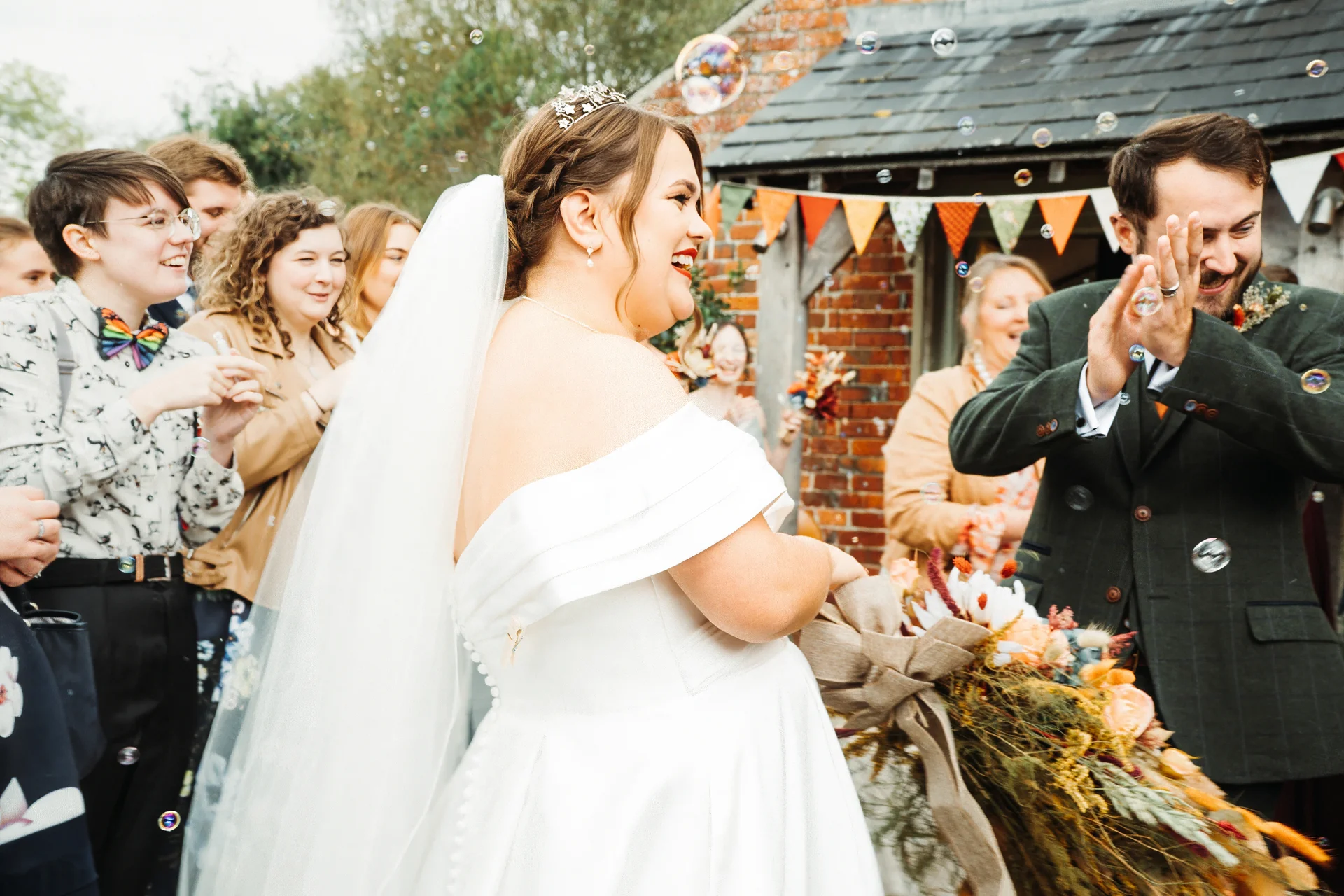 A joyful bride and groom laugh as bubbles float around them outside a rustic brick barn venue during their wedding celebration.