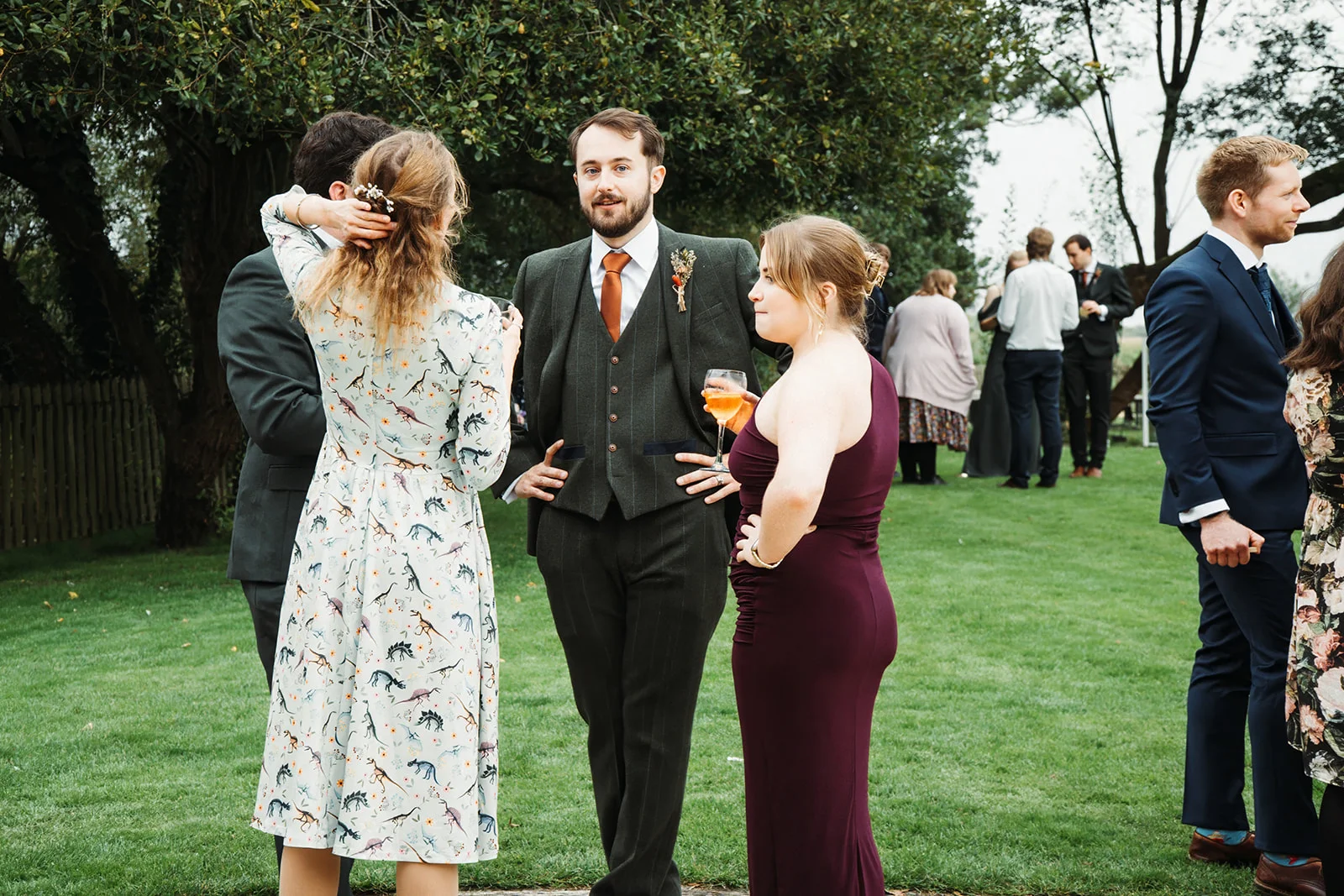 Wedding guests mingle on a grassy lawn during an outdoor drinks reception, with a groomsman in a dark green three-piece suit holding a glass of amber-coloured drink.