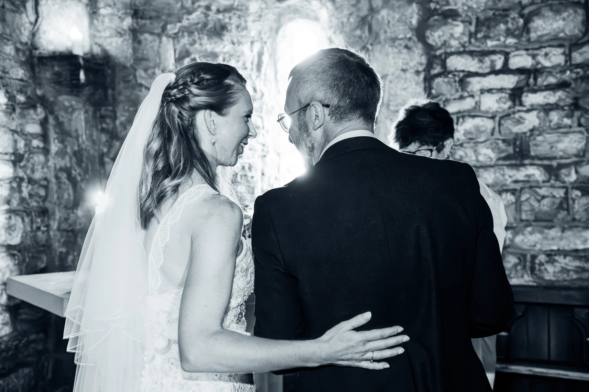 A newlywed couple embraces lovingly inside a historic stone venue, captured in a dramatic black and white photograph with backlit window light.
