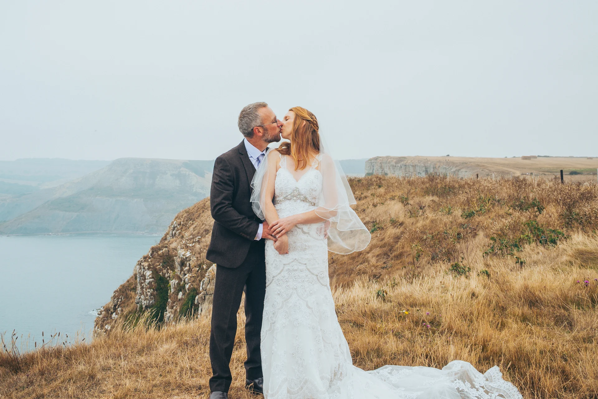 An older bride and groom share a tender kiss on a dramatic coastal clifftop against a backdrop of rolling hills and an overcast sky.