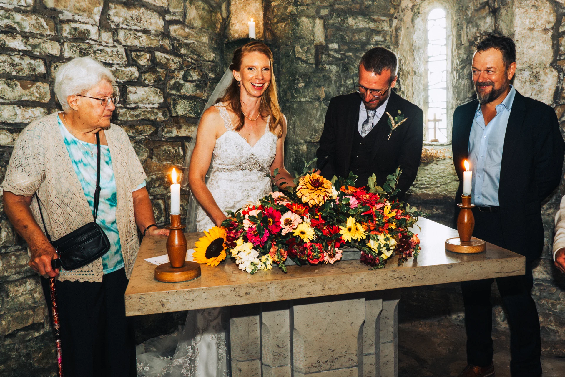 The bride and groom smile happily while signing the marriage register at a stone altar adorned with vibrant autumn flowers and burning candles, accompanied by family witnesses in an ancient stone church.