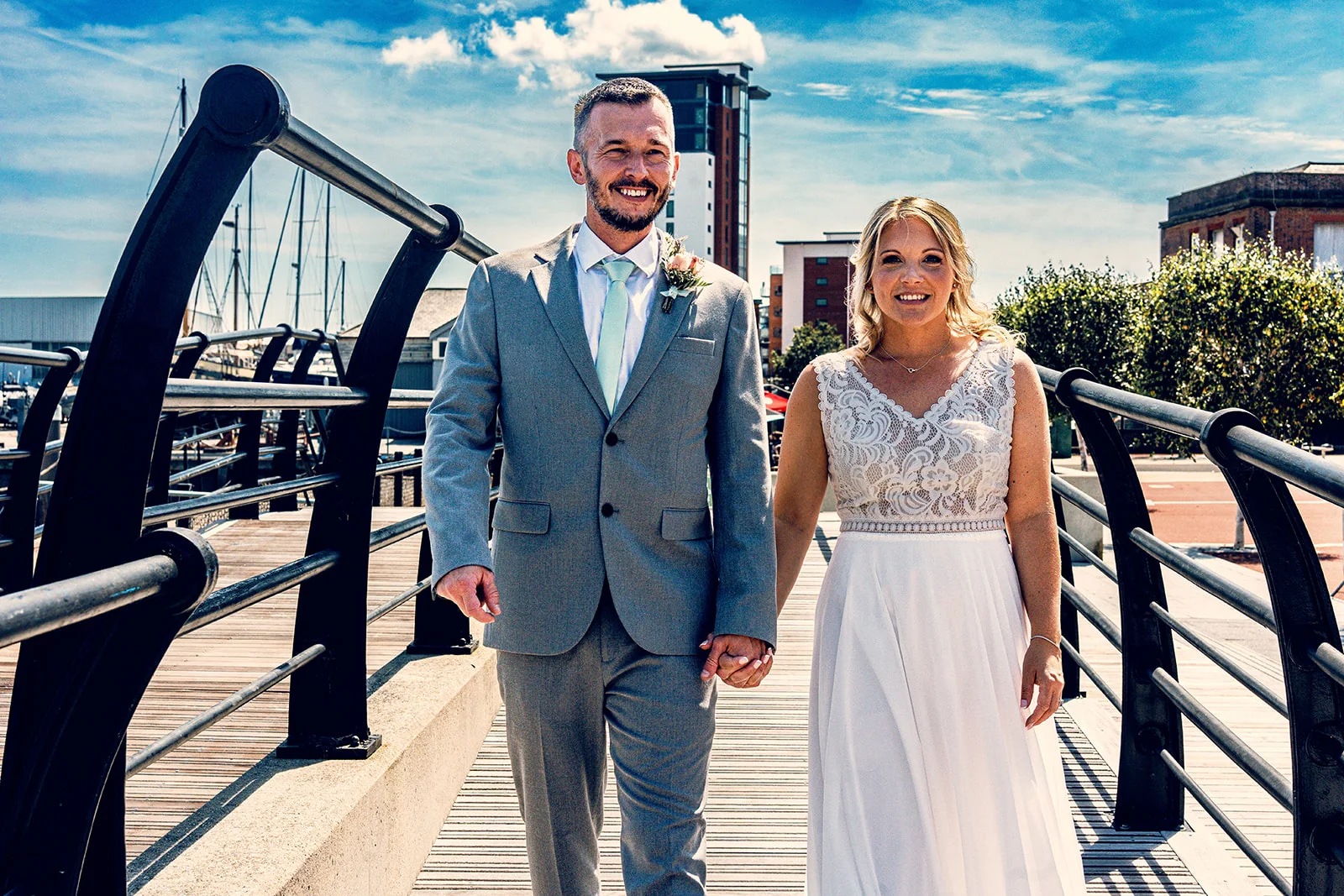 A smiling bride and groom holding hands while walking along a sunny harbour bridge during their city wedding portrait session.