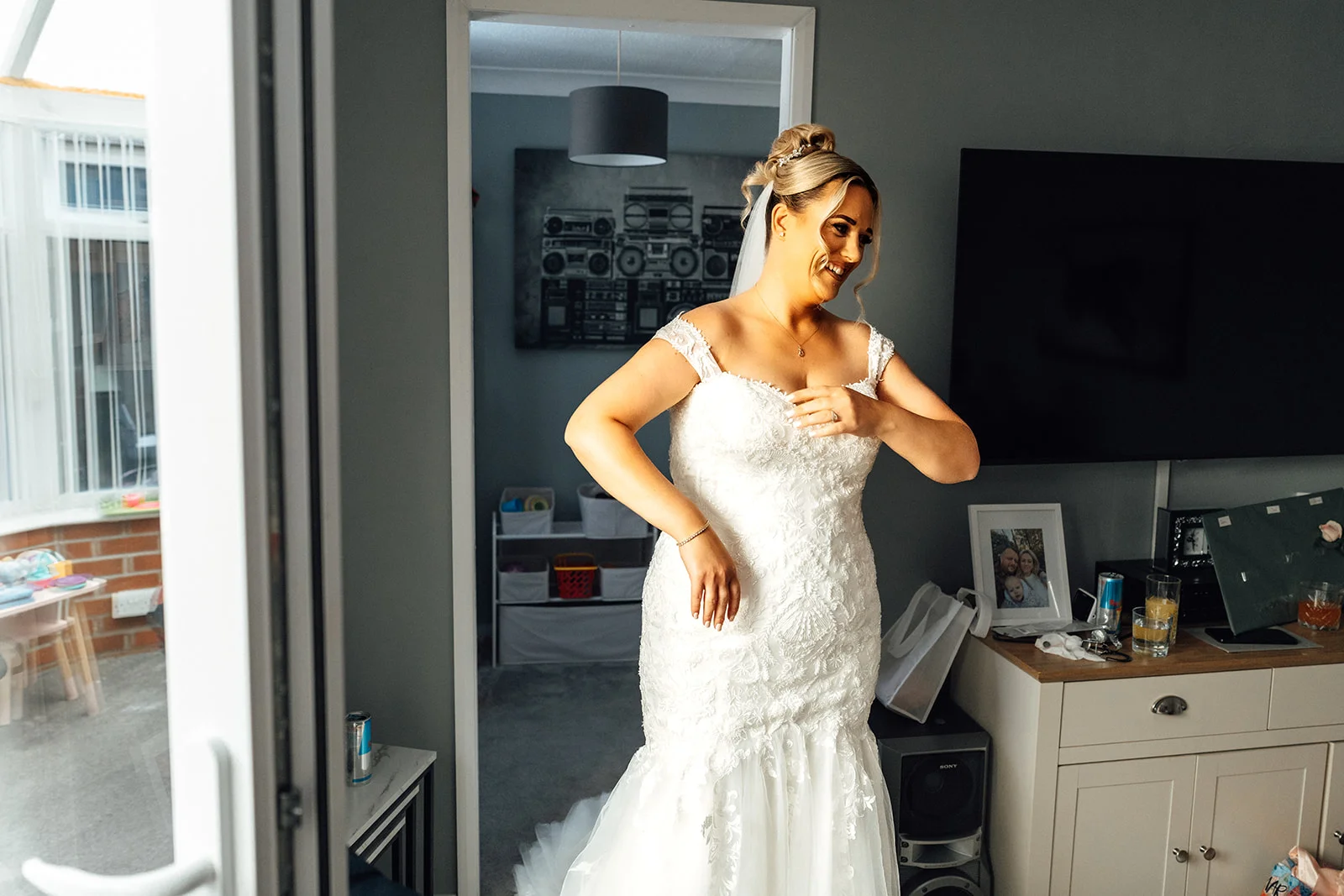 A smiling bride wearing an off-the-shoulder lace fishtail dress and veil stands in a room during bridal preparations.