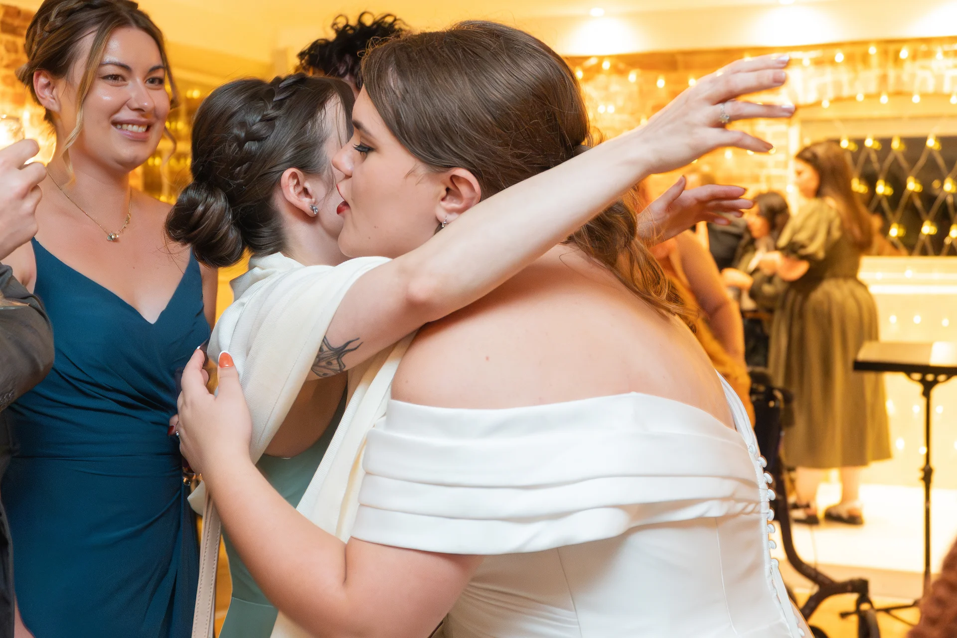 Two brides embrace tenderly during a wedding reception while guests mingle in the softly lit background.