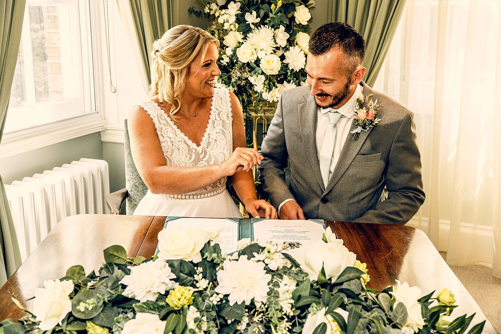 The bride and groom smile happily as they sign the wedding register behind a table adorned with white floral arrangements.