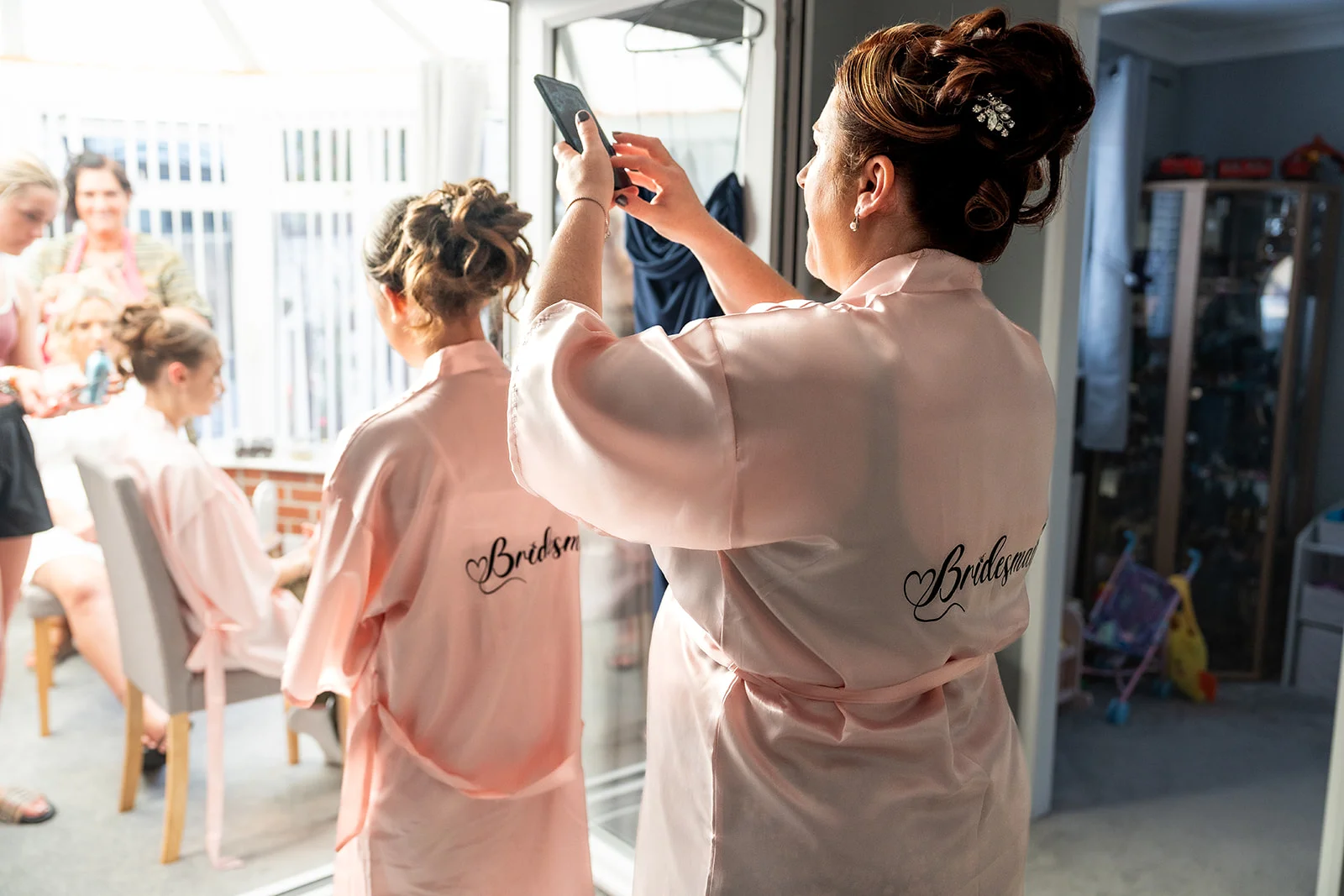 A bridesmaid wearing a personalised pink satin robe takes a photo on her phone while getting ready with friends on the morning of the wedding.