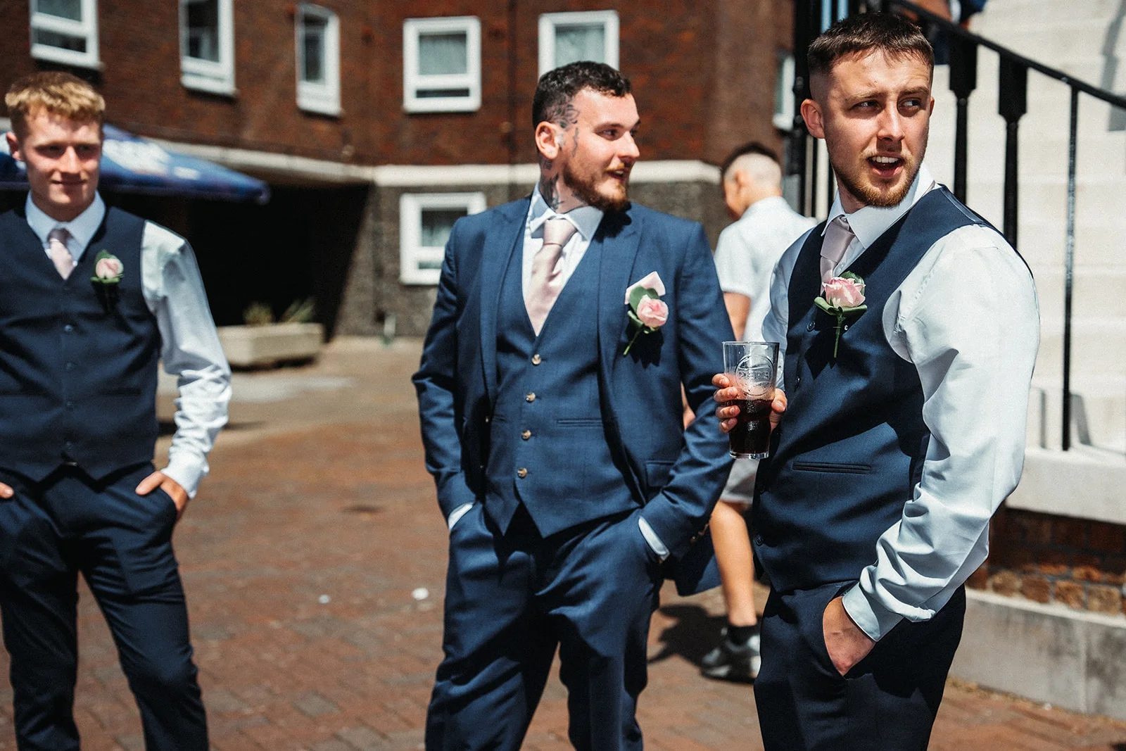 A group of groomsmen wearing matching navy three-piece suits and pink ties stand outdoors on a paved courtyard, chatting and holding a drink.
