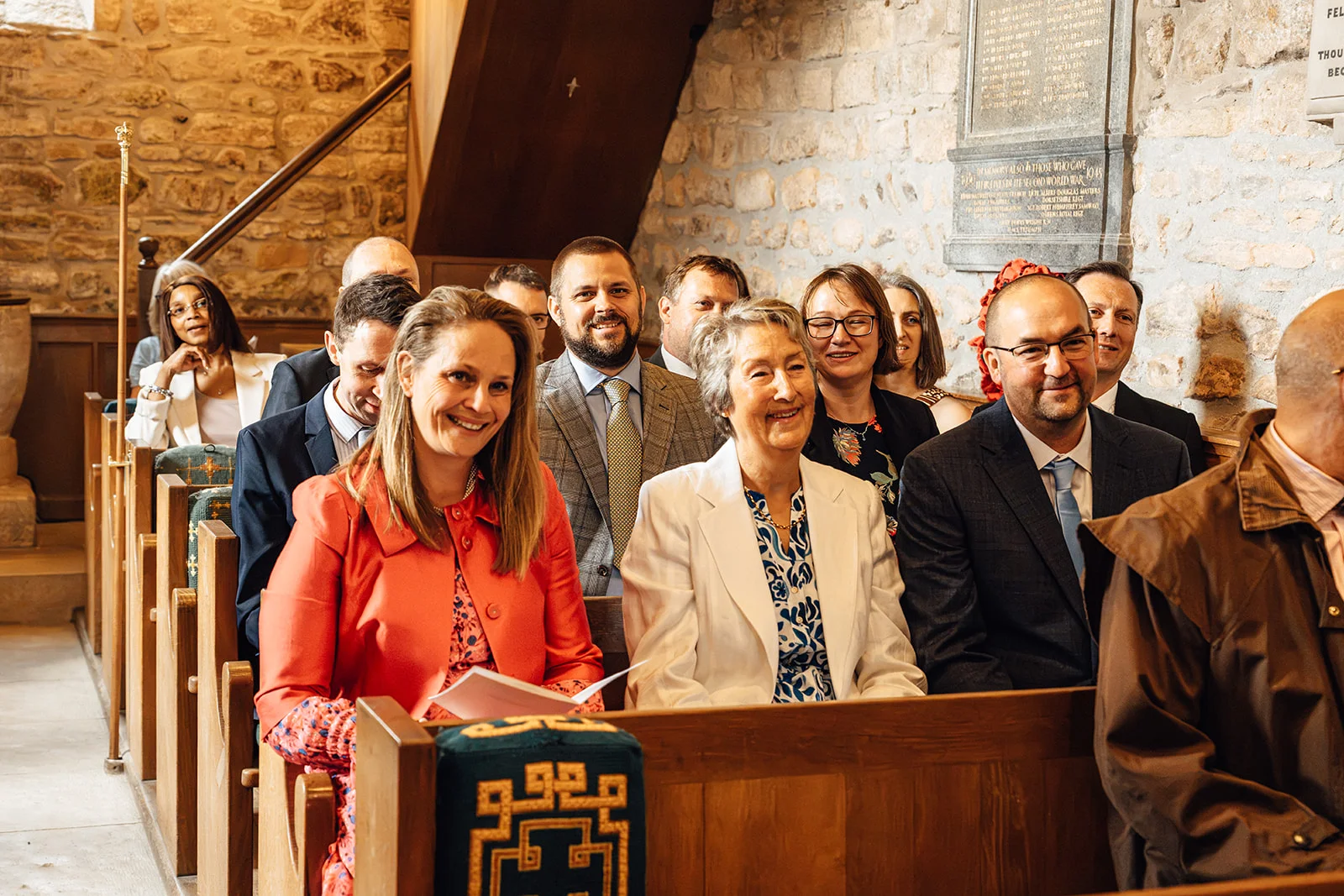 A group of smiling wedding guests seated in wooden pews inside a historic stone church during the ceremony.