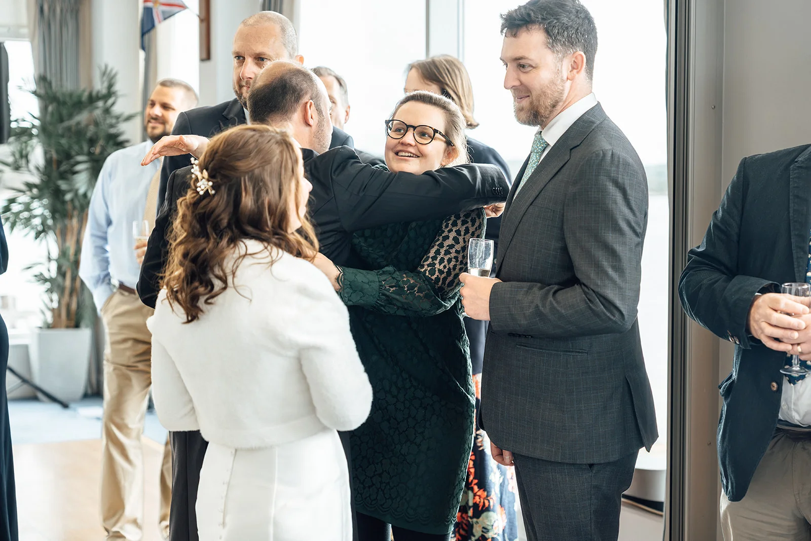 Wedding guests mingle and share joyful hugs and conversation during a bright drinks reception inside a modern venue.